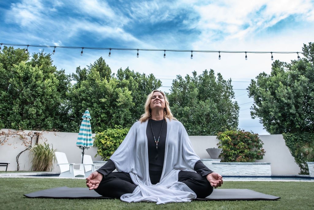 A woman practicing yoga outdoors on a black mat, sitting cross-legged with her hands resting on her knees, eyes closed, appearing relaxed. She is surrounded by trees, a swimming pool, outdoor furniture, and a blue sky with some clouds.