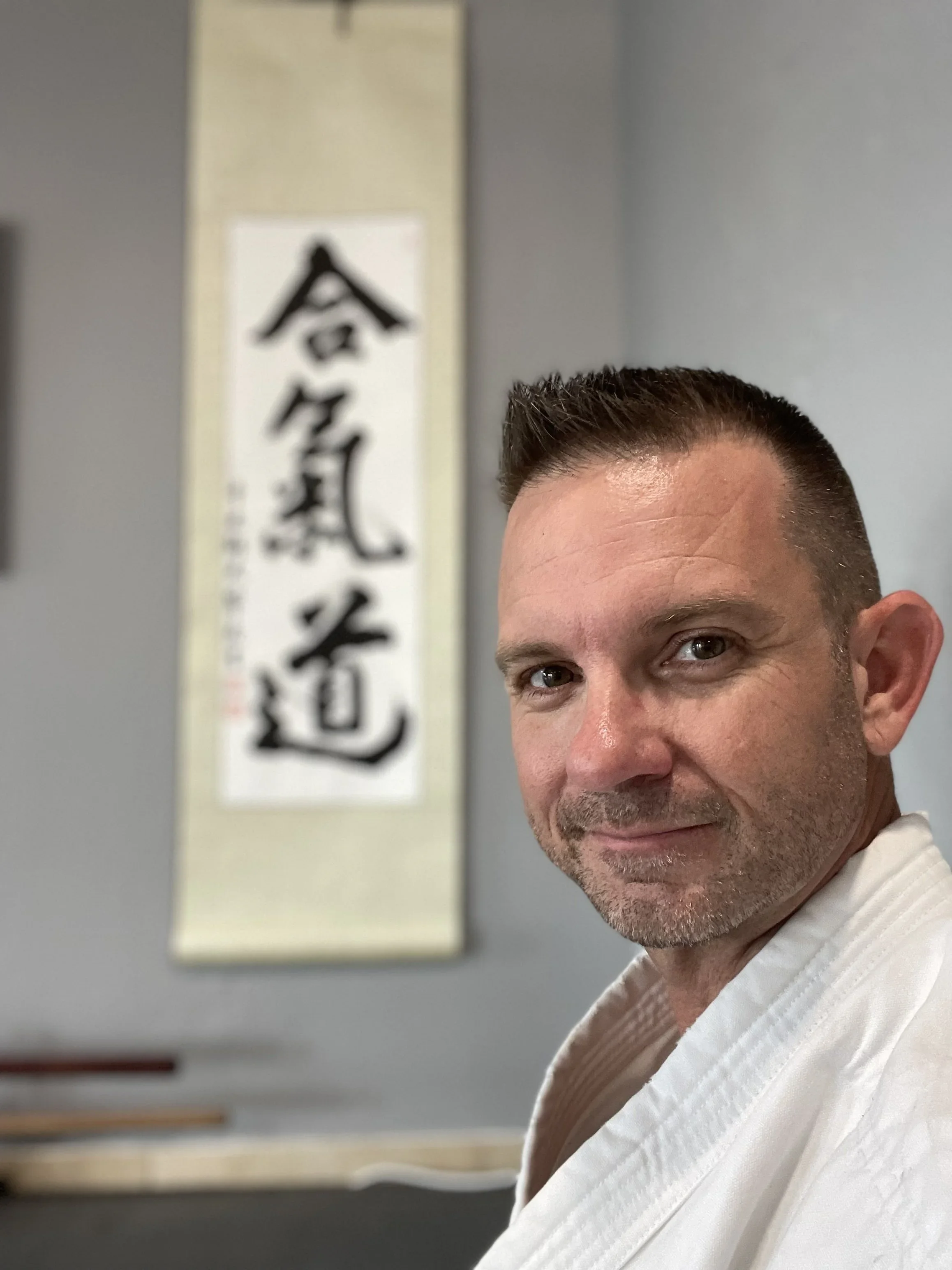A man with short dark hair and a beard smiling in a martial arts uniform inside a dojo with Japanese calligraphy on a wall in the background.