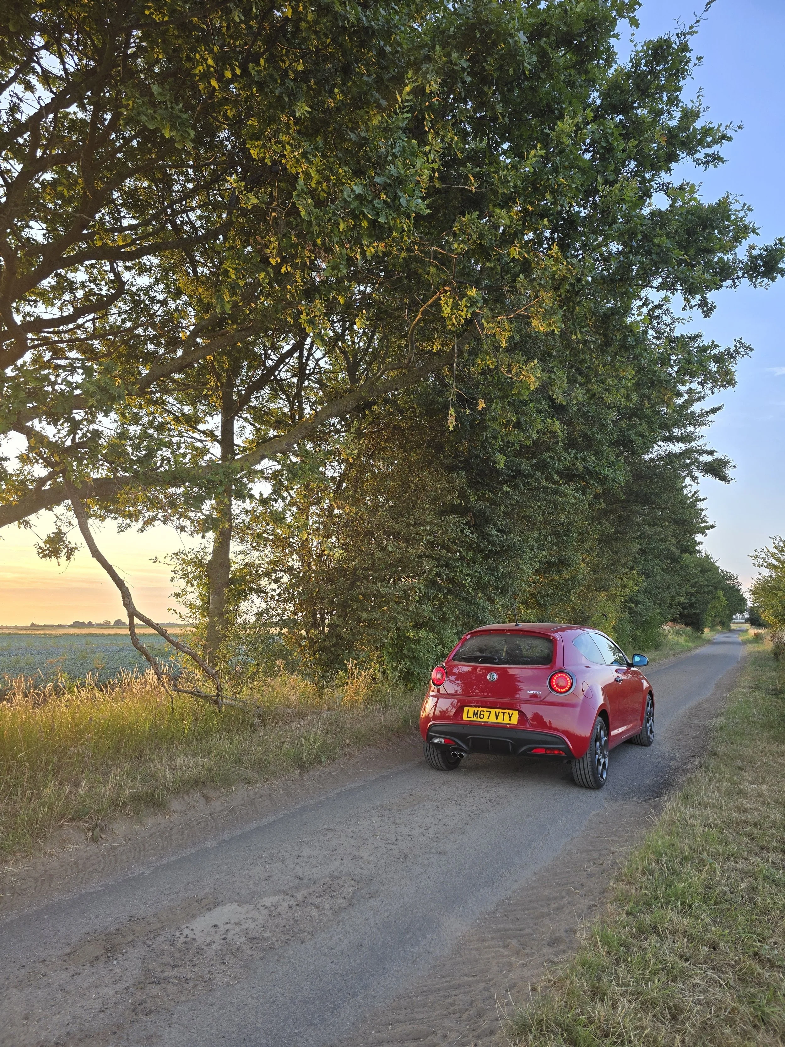 Red Alfa Romeo Mito Veloce parked on a narrow country road against the backdrop of a sunset.