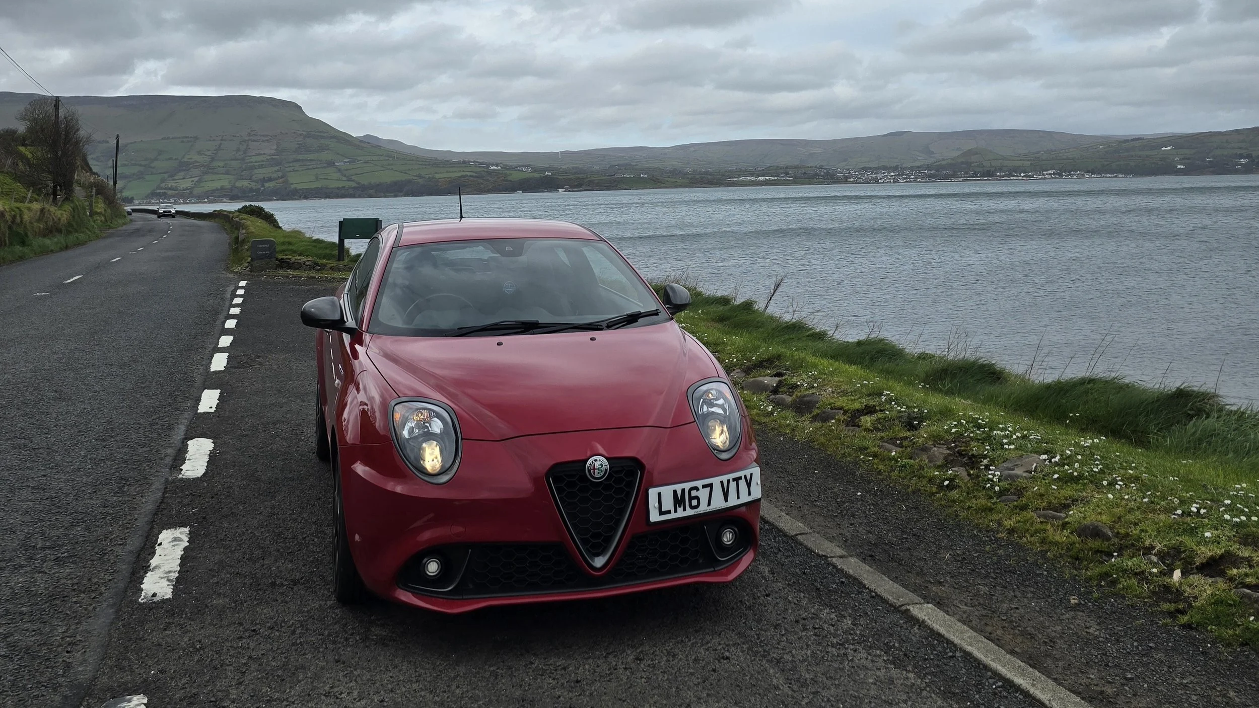 Red Alfa Romeo Mito parked by the side of a road on a scenic coastline on a grey, overcast day.