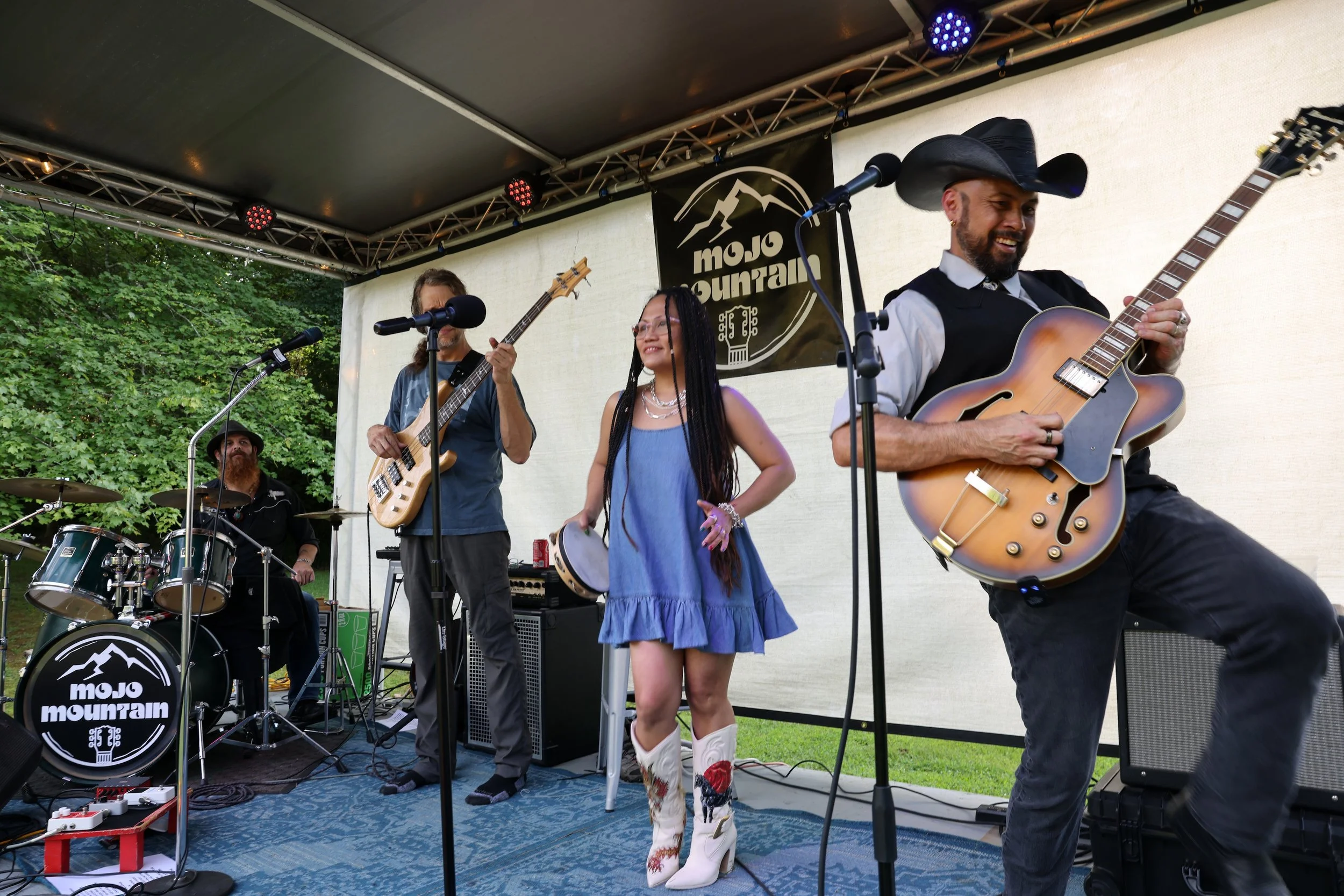 A live outdoor music performance featuring a band, including a drummer, a bassist, a woman singer, and a guitarist, on a stage with a sign that reads 'mojo mountain'. The guitarist is wearing a cowboy hat and jeans, and the female singer is wearing a blue dress with cowboy boots. The stage is set against a backdrop of green trees.