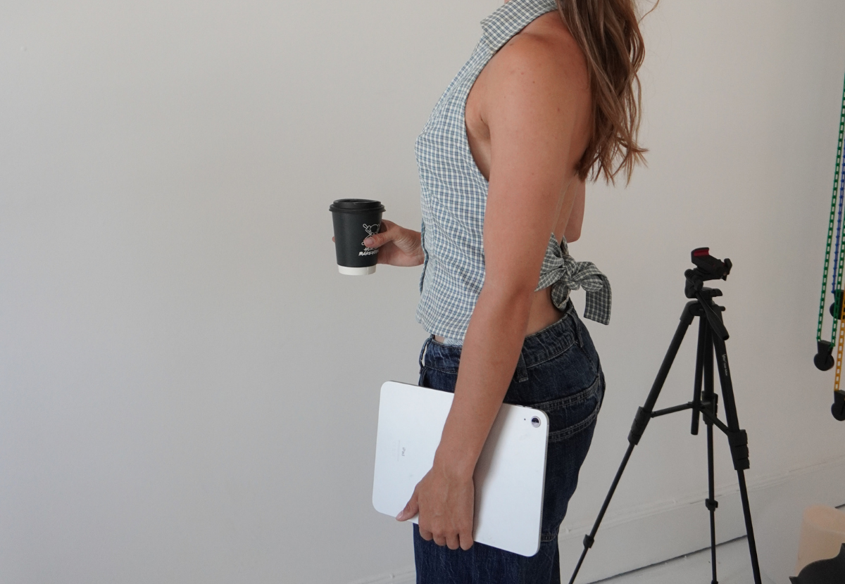 Woman with long wavy hair wearing a blue and white checkered sleeveless top tied at the waist, holding a black coffee cup in her right hand and a closed white laptop in her left hand, standing next to a tripod in a room with a plain white wall.