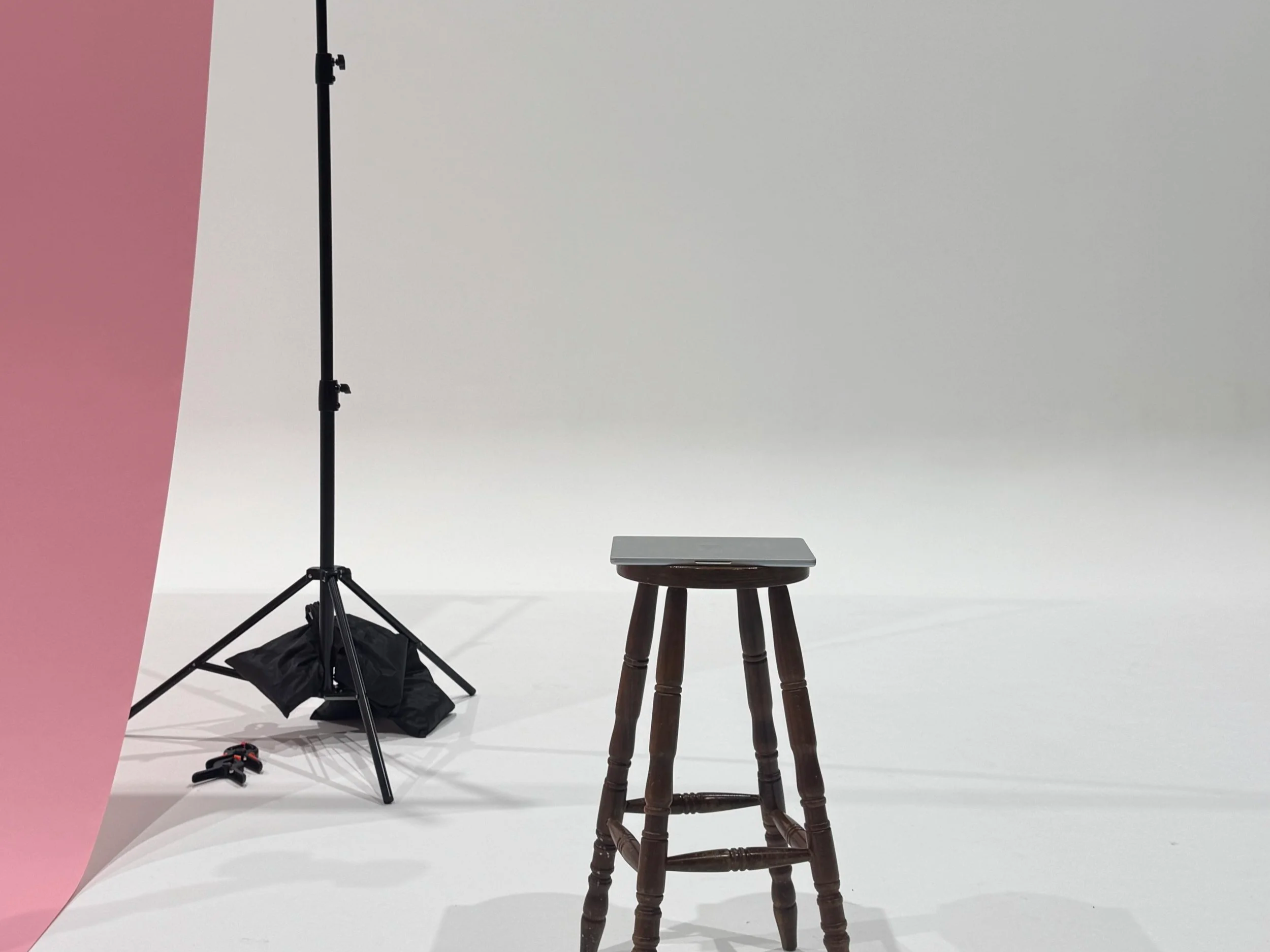 Photography setup with a black tripod stand, a stool with a closed silver laptop, a pink backdrop on the left, and a plain white background.