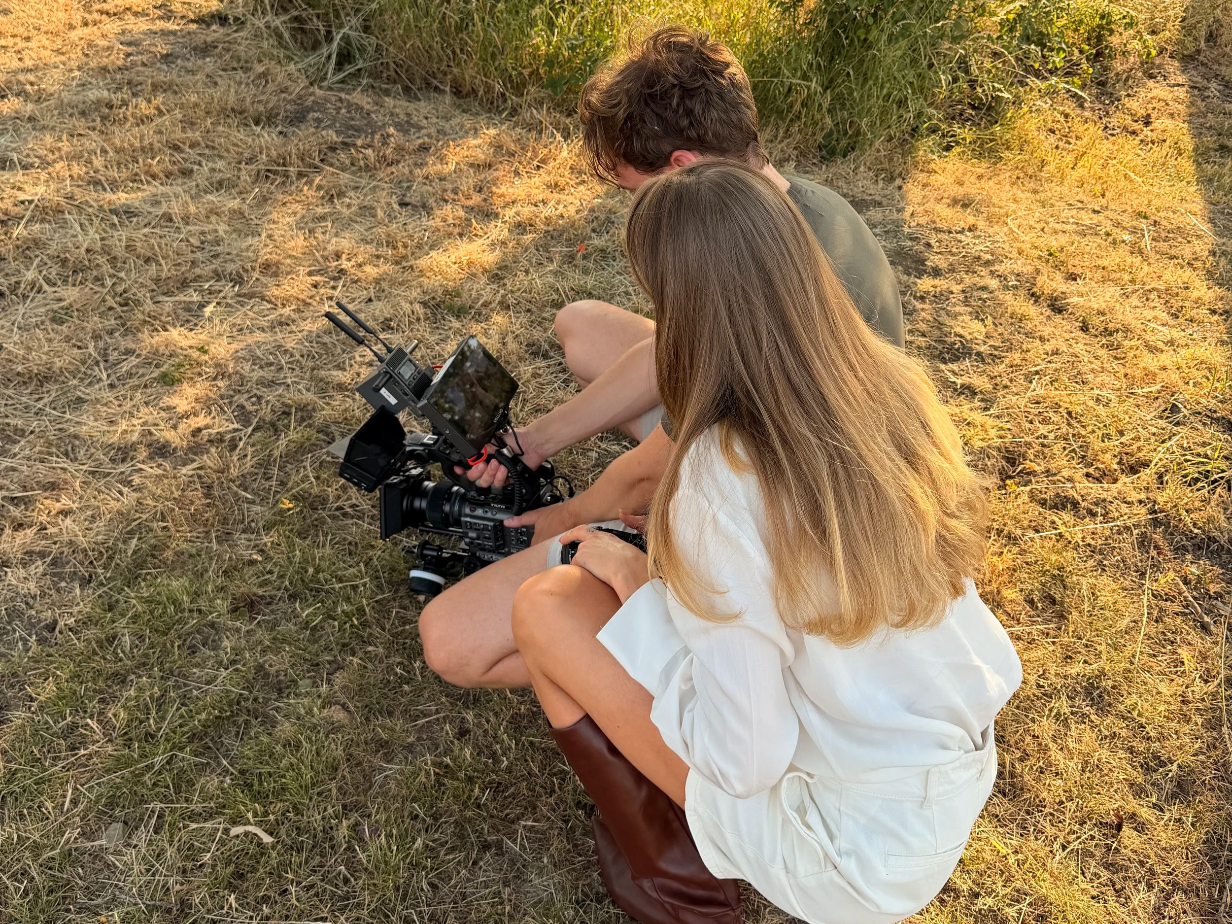 Two people sitting on dry grass in a field during sunset. One person, with long blonde hair, in a white shirt, is holding a professional camera. The other person, with short brown hair, wearing a green shirt, is looking at the camera's screen.