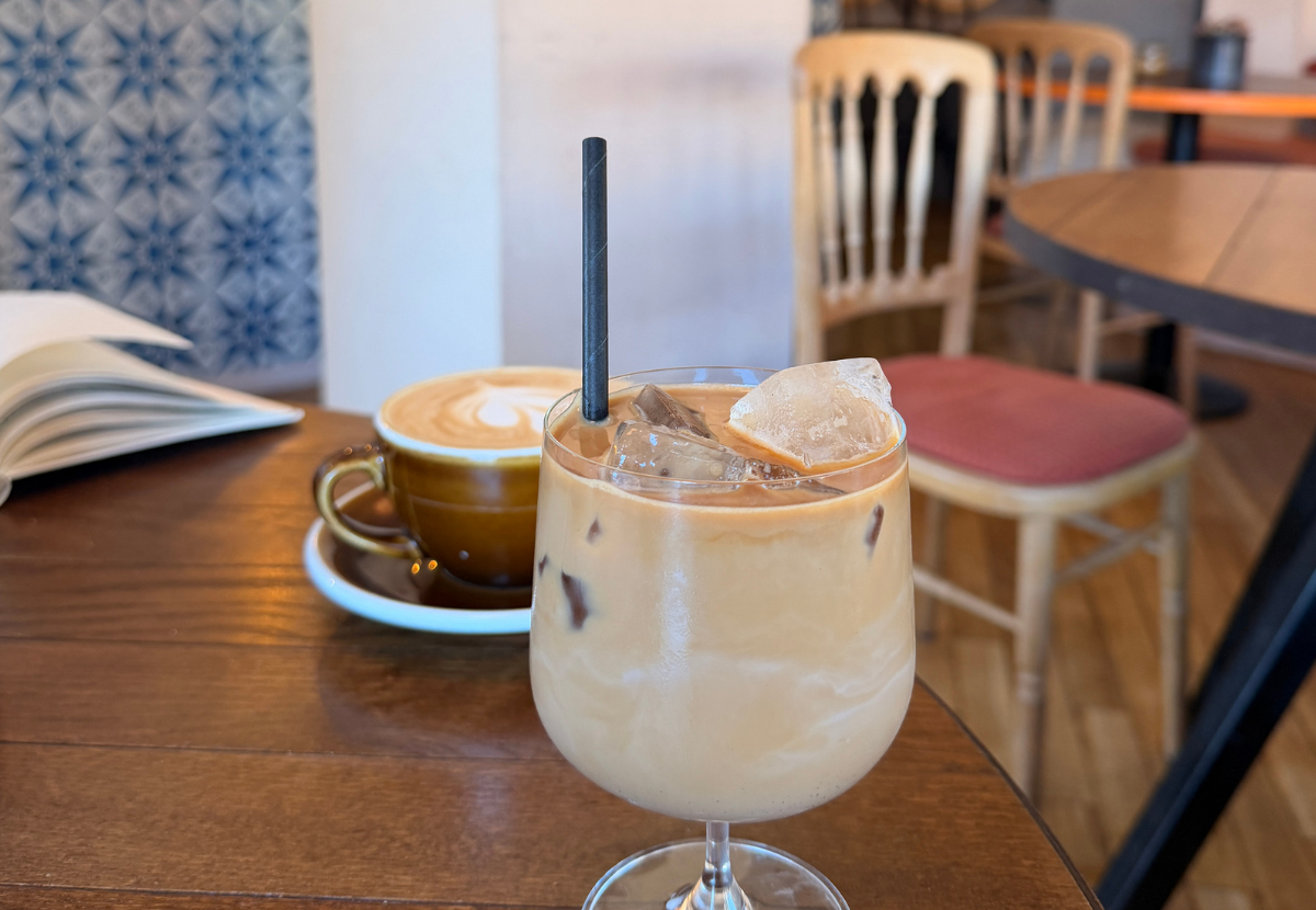 An iced coffee drink with ice cubes and a straw on a wooden table in a café, with a cup of latte and an open book in the background.