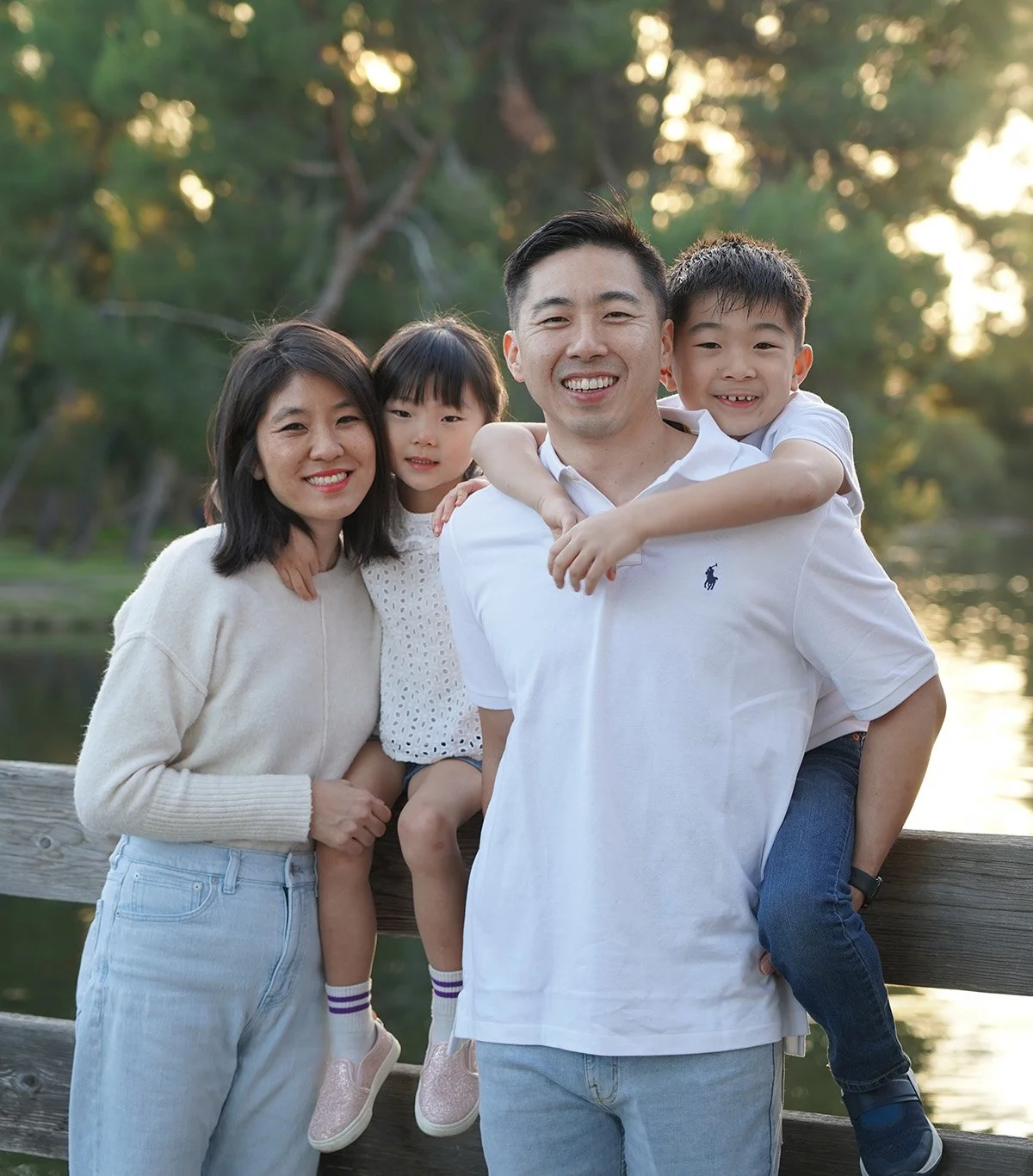 A smiling family (Albert and Elisa + kids) of four outdoors with a lake and trees in the background during sunset. The mother stands on the left, the father holds a young boy on his back, and a young girl is sitting on a wooden railing between them.