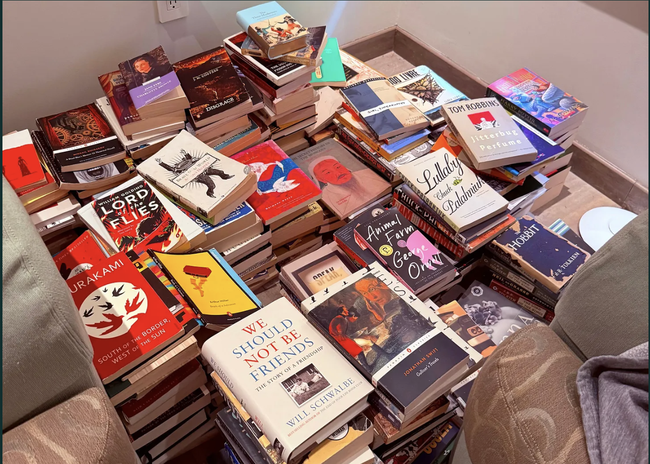 A large collection of books on tables and the floor in a cluttered arrangement, surrounded by two chairs and a corner of a room with a electrical outlet on the wall.