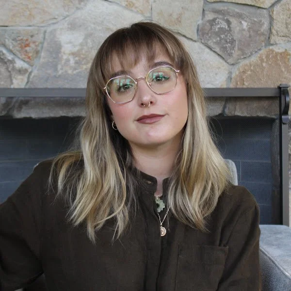 Young woman with glasses and blonde hair sitting in front of a fireplace