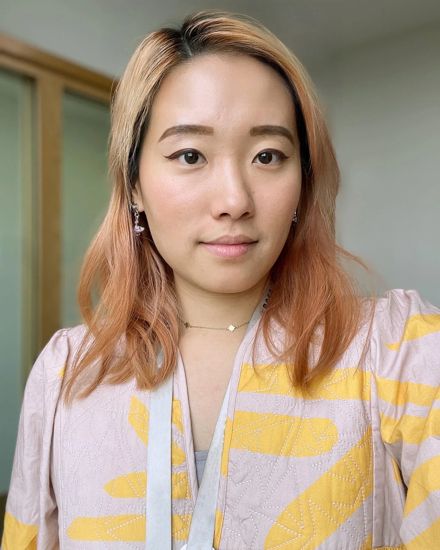 A woman with hair dyed in a reddish-orange hue, smiling softly at the camera. She is wearing a light pink top with yellow abstract patterns, silver earrings, a delicate star necklace, and light makeup with winged eyeliner. The background includes a light-colored wall and a wooden-framed sliding door.