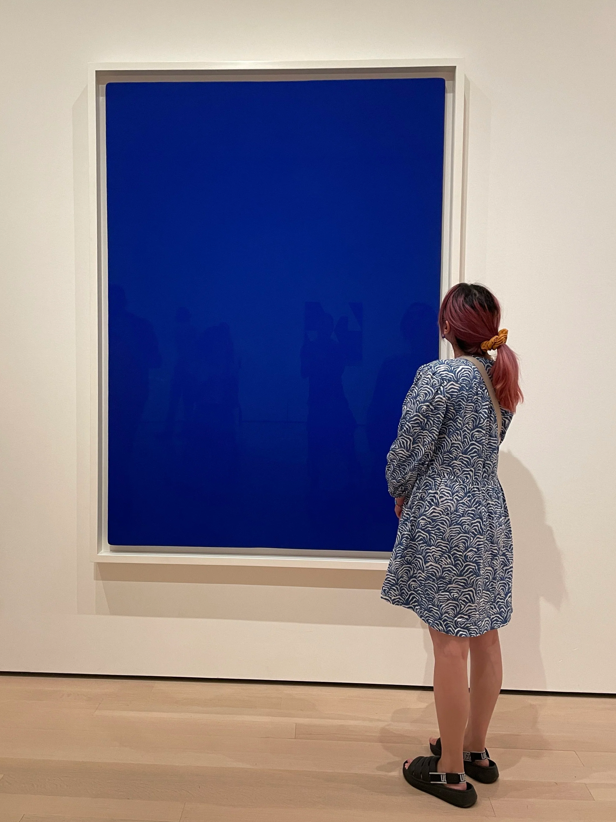 A person with pink hair tied with a yellow scrunchie wearing a patterned blue and white dress, standing in front of a large, framed blue artwork in a gallery.