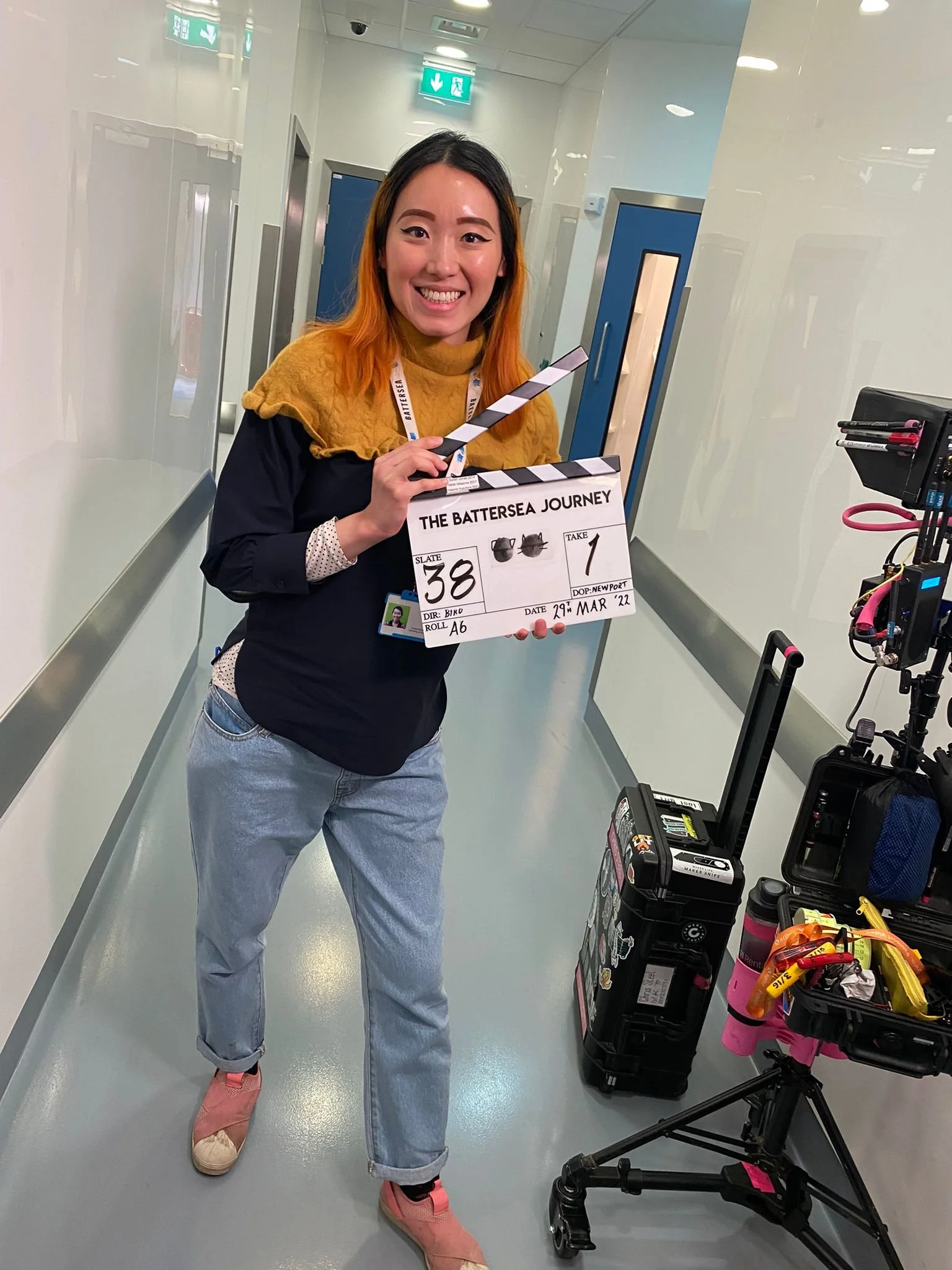 A woman with shoulder-length orange hair and winged eyeliner holding a clapperboard that reads 'The Battersea Journey' stands in a corridor, smiling at the camera. She is wearing a black top, light blue jeans, and pink sneakers. There are equipment and tools on a cart next to her, and emergency exit signs are visible on the ceiling.