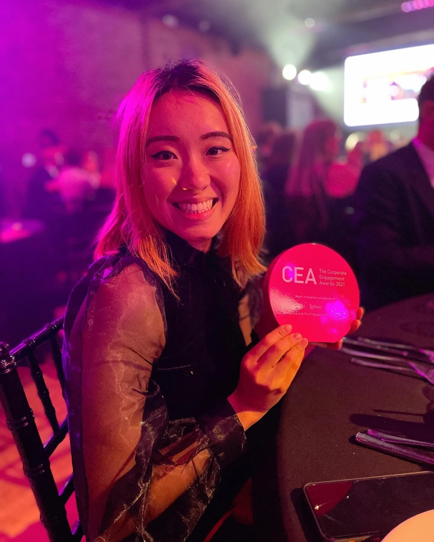 A woman with short orange hair smiling and holding a pink award at an event, surrounded by other people in a dimly lit venue with colorful lighting.