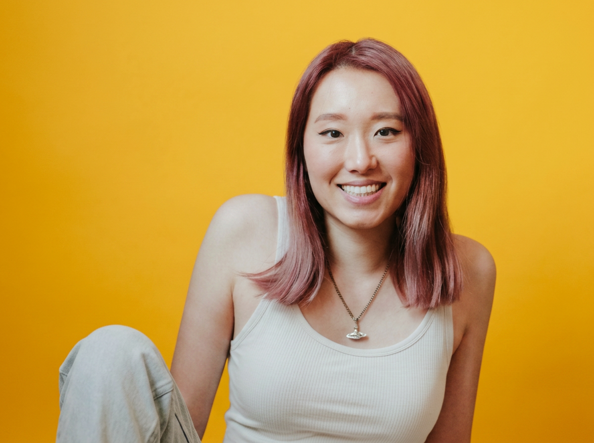 A young woman with shoulder-length pink hair, smiling, wearing a white tank top and a necklace, sitting against a bright yellow background.
