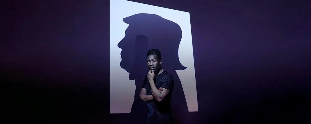 Theatre production photo of a man in a black t-shirt standing with arms crossed in front of a large illuminated screen showing the shadow silhouette of a man's head and hair.