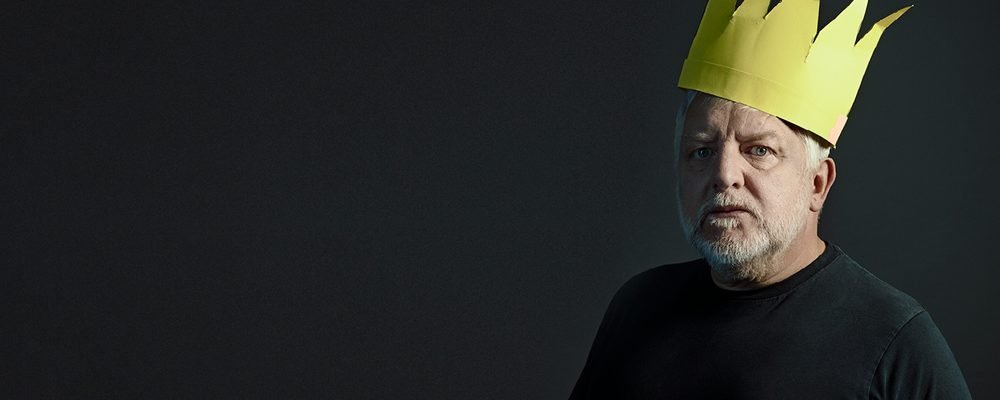 Theatre production photo of an older man with grey hair and beard wearing a paper crown and plain black t-shirt, staring solemnly at the camera against a dark background.