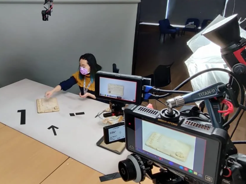 A woman wearing a purple face mask is sitting at a table with numbered markers, a camera setup is filming her, and she is reaching for a small wooden object on the table. The room has a modern design with chairs and tables in the background, and lighting appears to be controlled for filming.