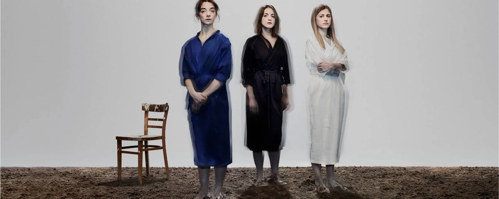 Theatre production photo of three women standing barefoot on bare earth in matching but differently coloured robes (blue, black, and white), with an old wooden chair beside them, against a plain grey backdrop.