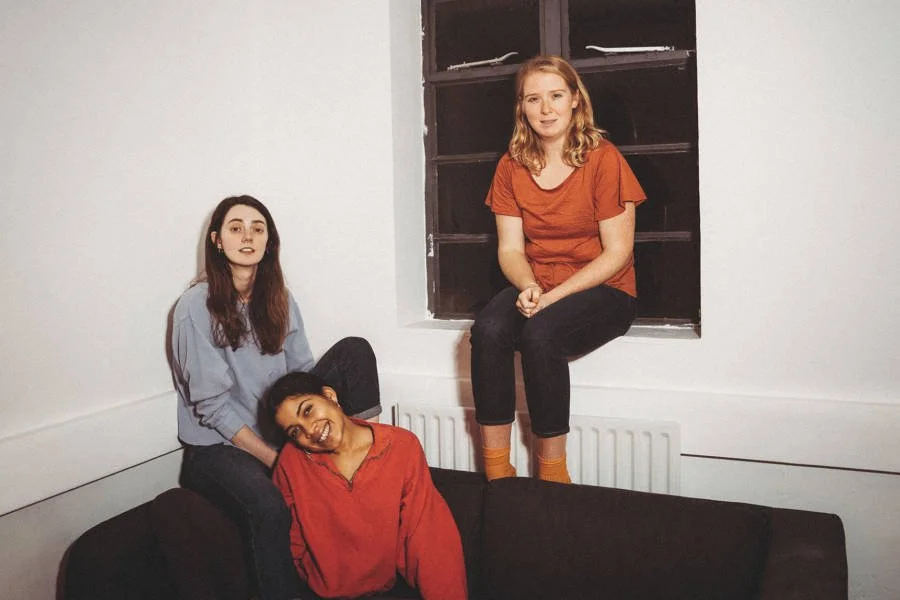 Promotional photo of three young women in a bare room with a dark-paned window, one perched on the windowsill and two sitting together on a brown sofa below, all looking toward the camera.