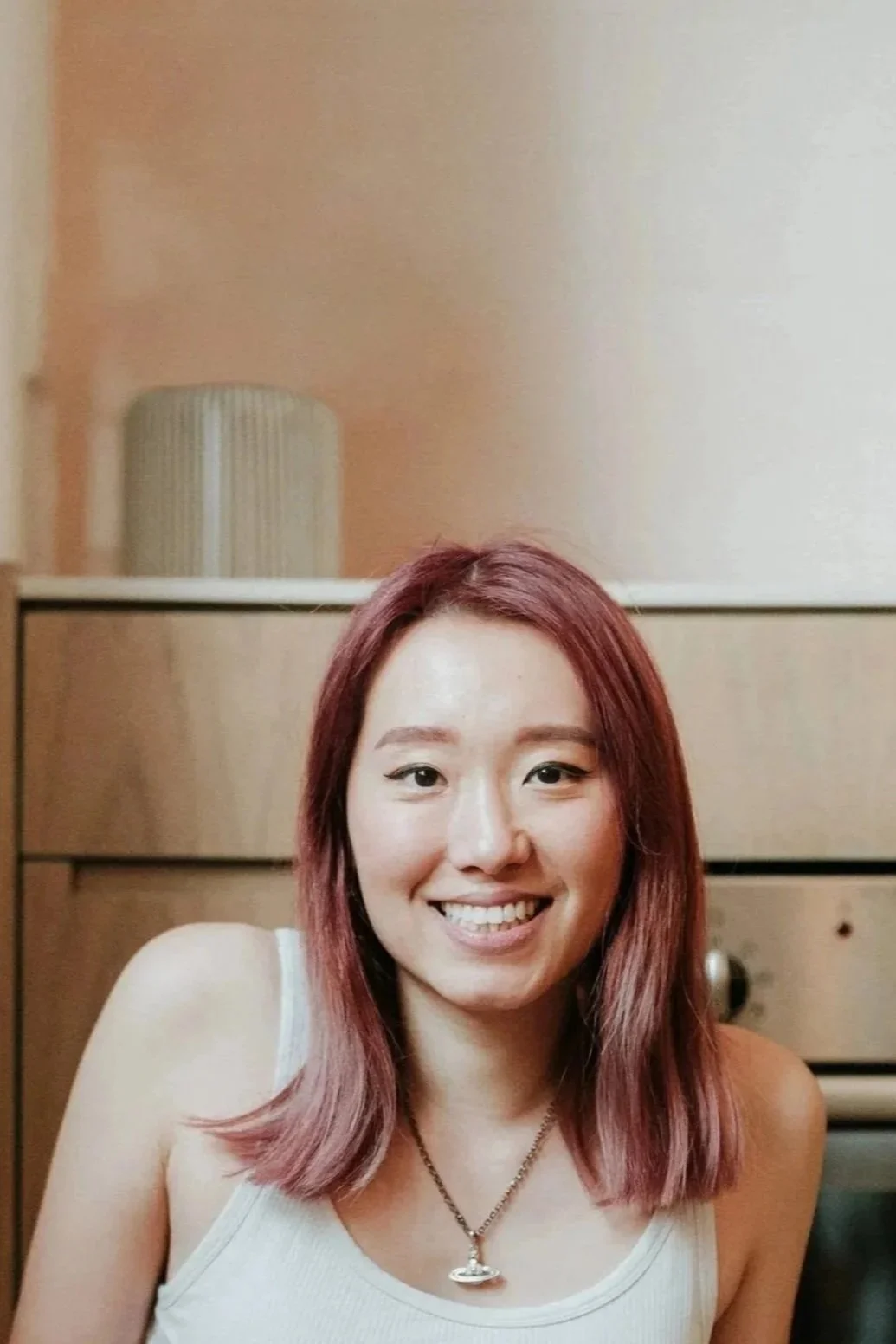 A woman with shoulder-length pink hair smiling, wearing a white tank top and a necklace with a small pendant, sitting in front of a wooden dresser and a beige wall.