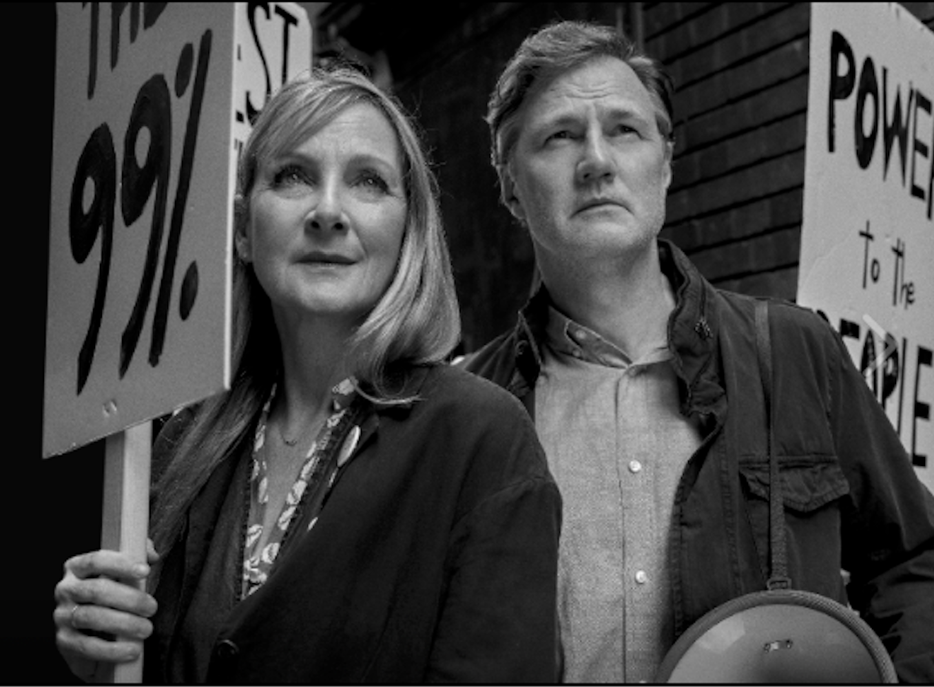 Black-and-white promotional photo of a woman and a man standing close together outdoors, the woman holding a protest sign reading "THE 99%" and the man holding a megaphone, with another sign reading "POWER TO THE PEOPLE" visible behind them.