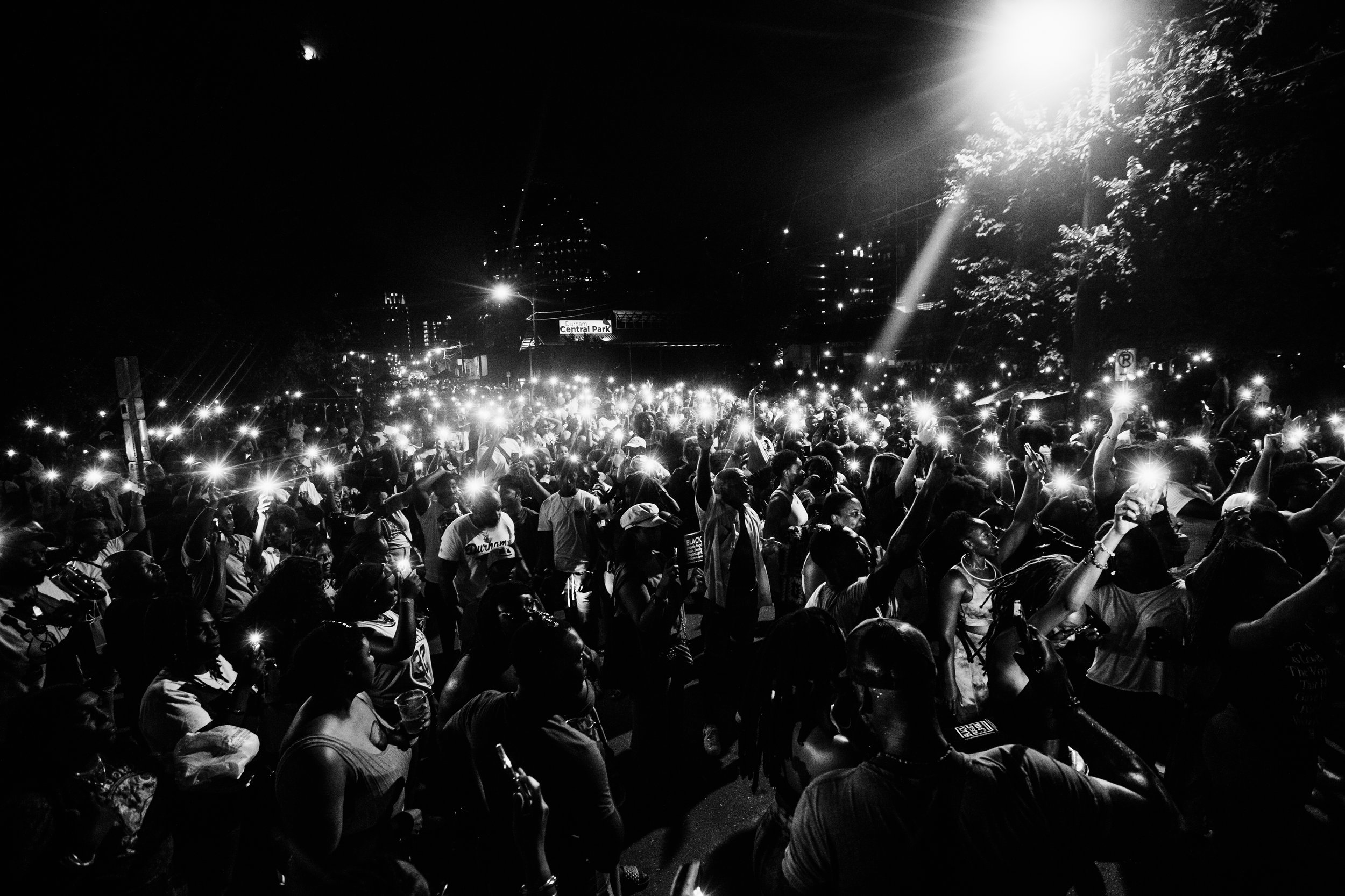 A large crowd of people at night holding up illuminated phones, gathered outdoors with trees and city buildings in the background.