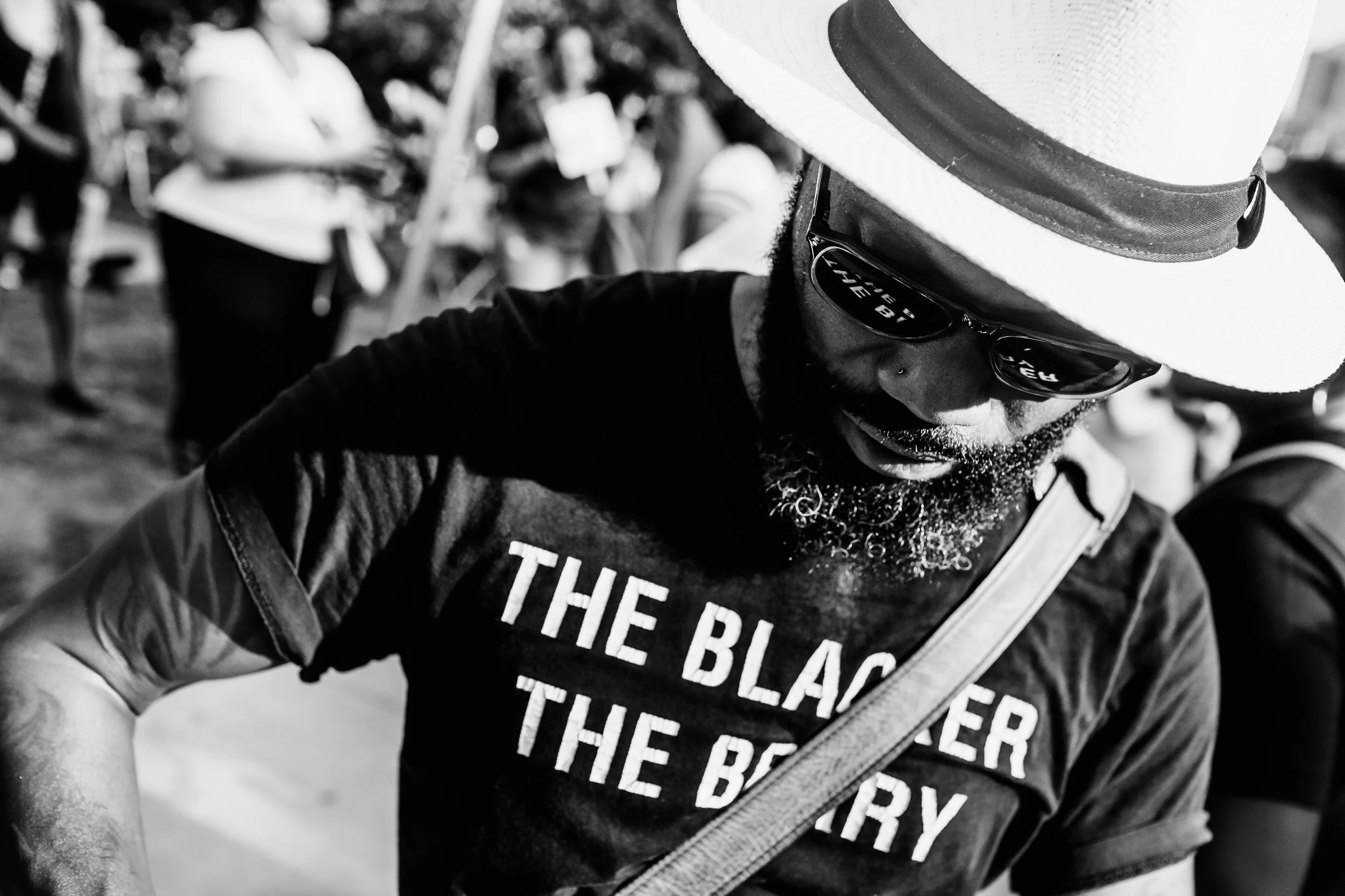 A person wearing a straw cowboy hat, sunglasses, and a black T-shirt with white text, leaning down while surrounded by others in an outdoor setting.