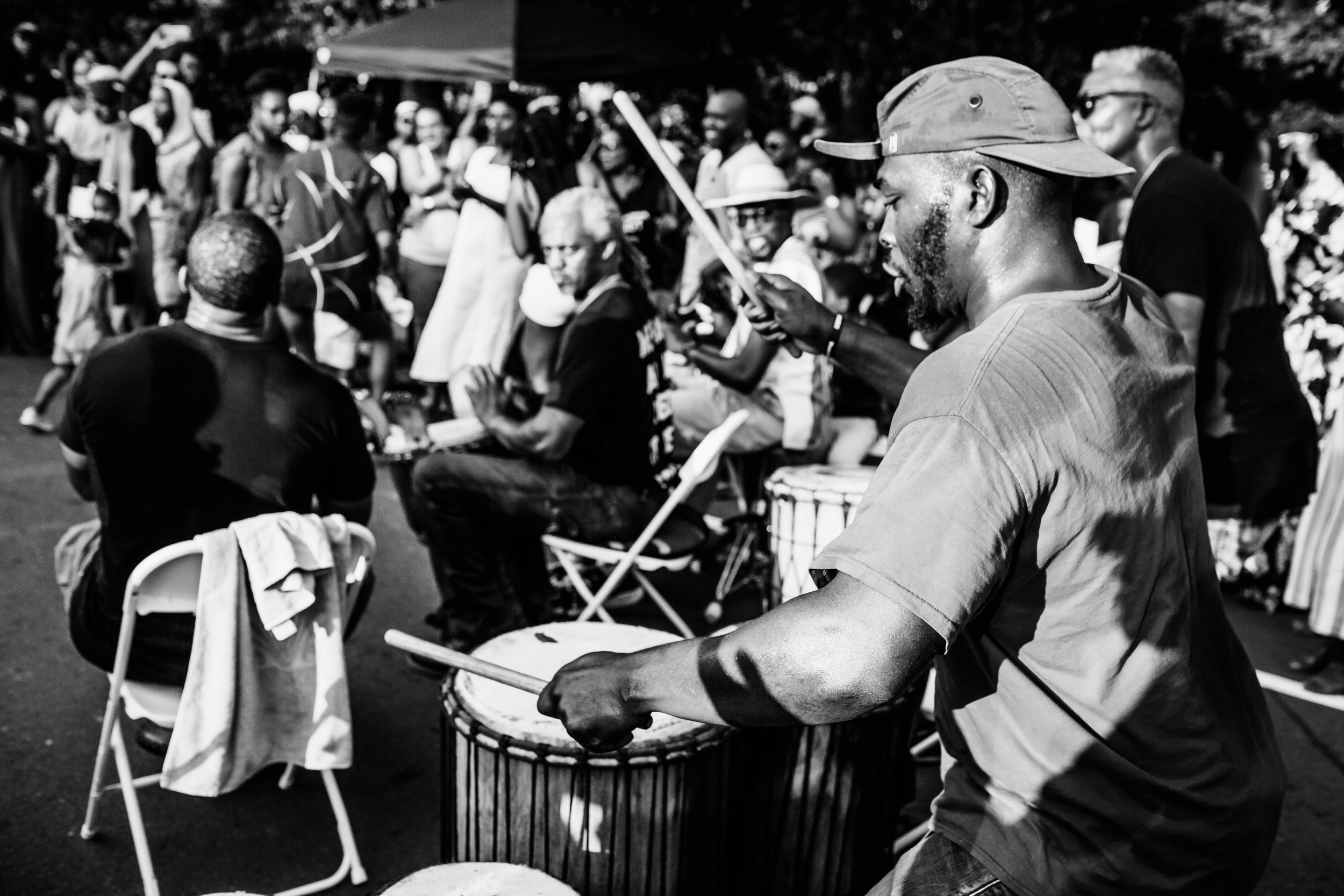 A black and white photo of a man playing a drum outdoors, surrounded by a crowd of people at the Black August in the Park event in Durham, North Carolina, photographed by Durham and Raleigh-based photographer, Dare Kumolu-Johnson.