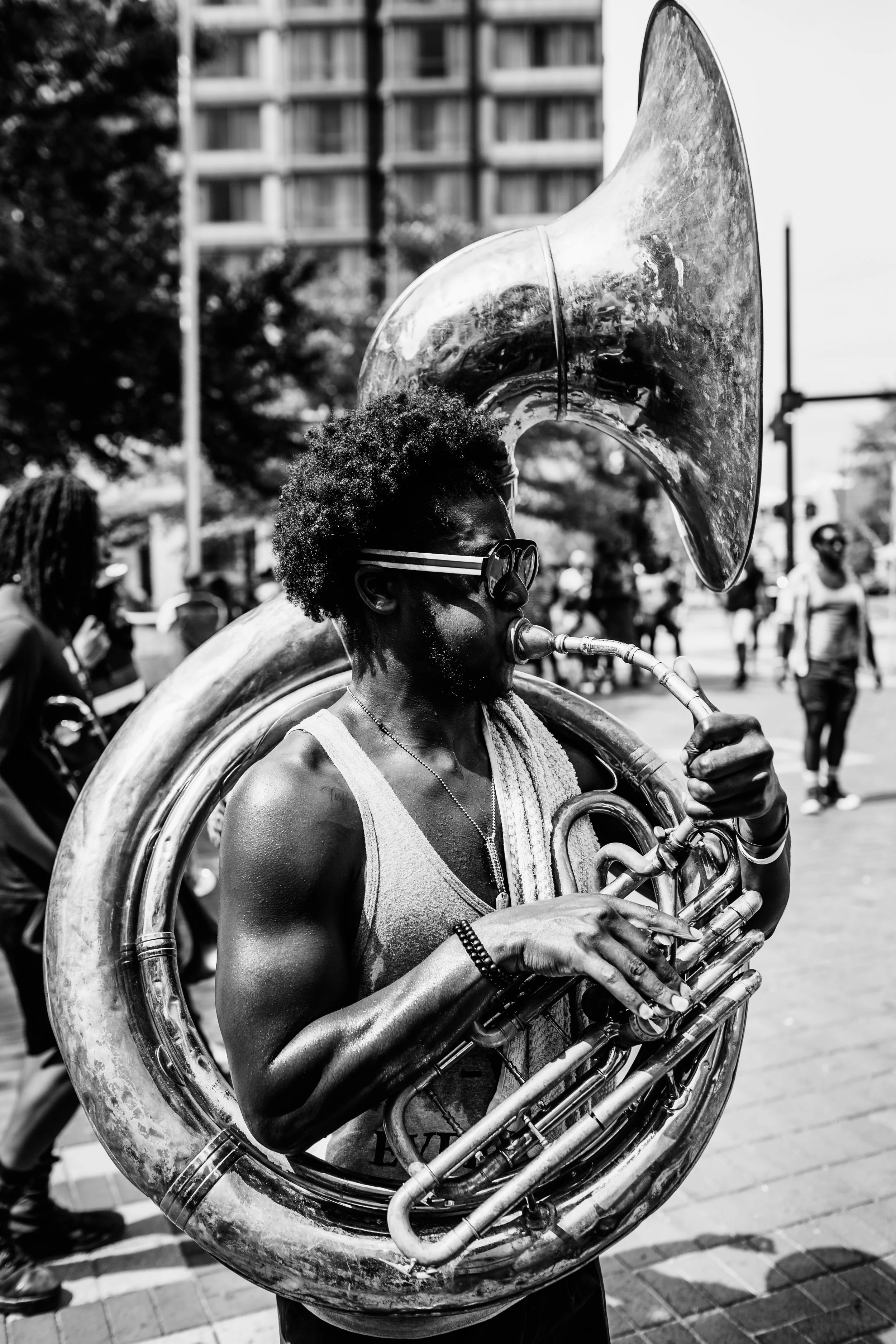 A Black man with curly hair wearing sunglasses playing a sousaphone during a street parade or gathering. Background shows trees, a building, and several other people.