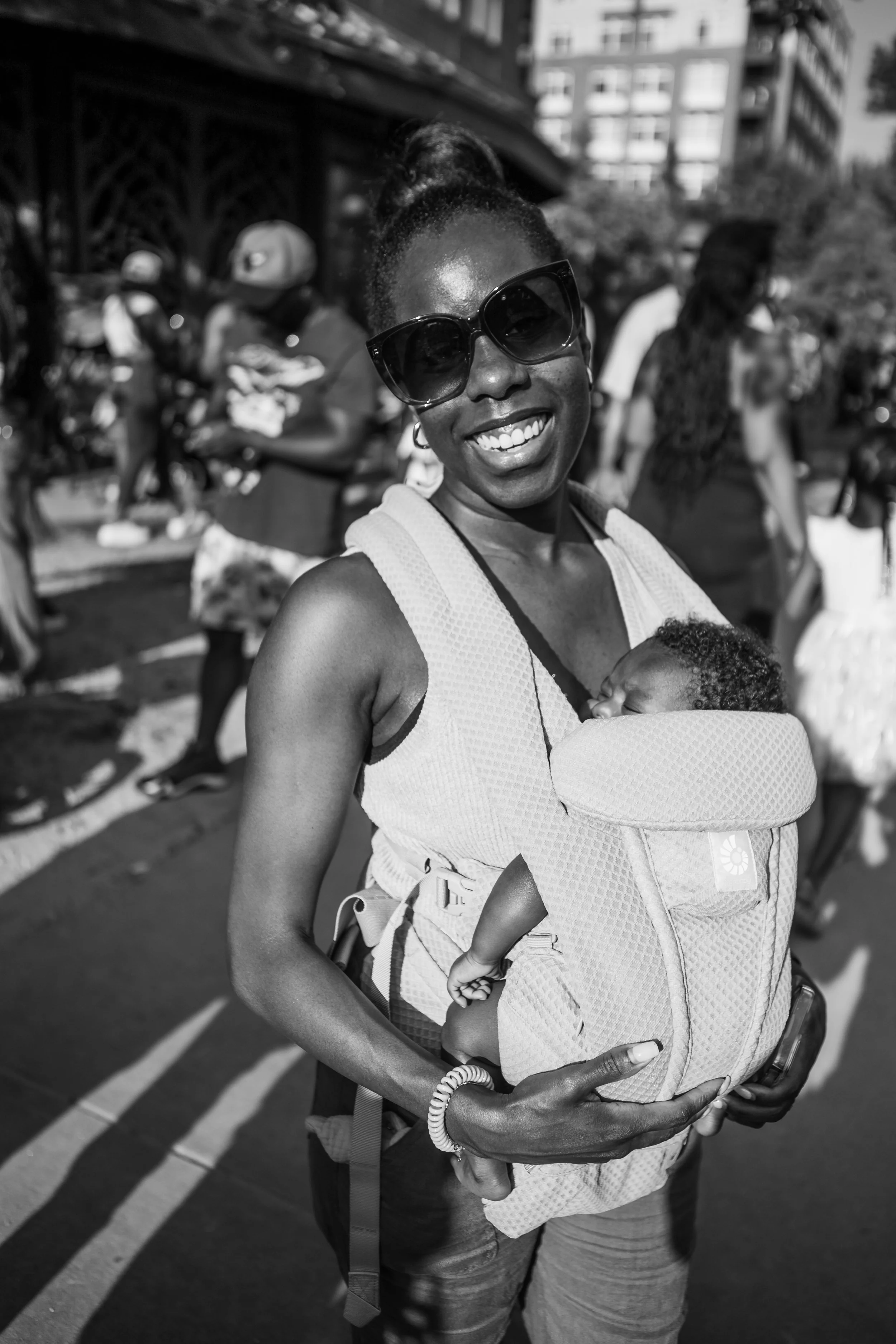 Happy woman wearing sunglasses and a tank top carrying a baby in a baby carrier, outdoors during daytime with a blurred background of people and buildings