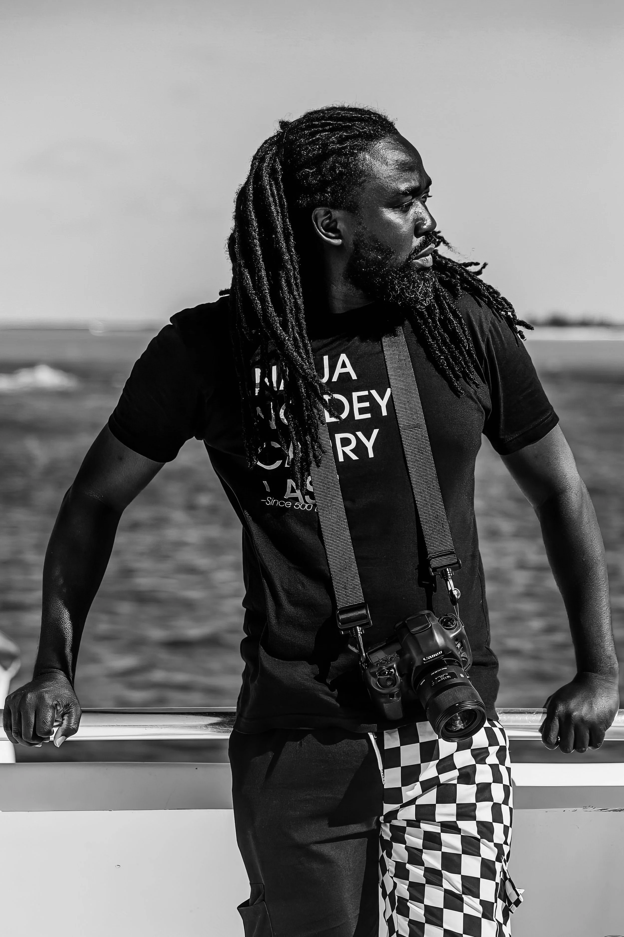 A man with dreadlocks and a beard standing on a boat holding a railing, with water in the background. He is wearing a black graphic t-shirt, checkered shorts, and has a camera hanging from his neck.