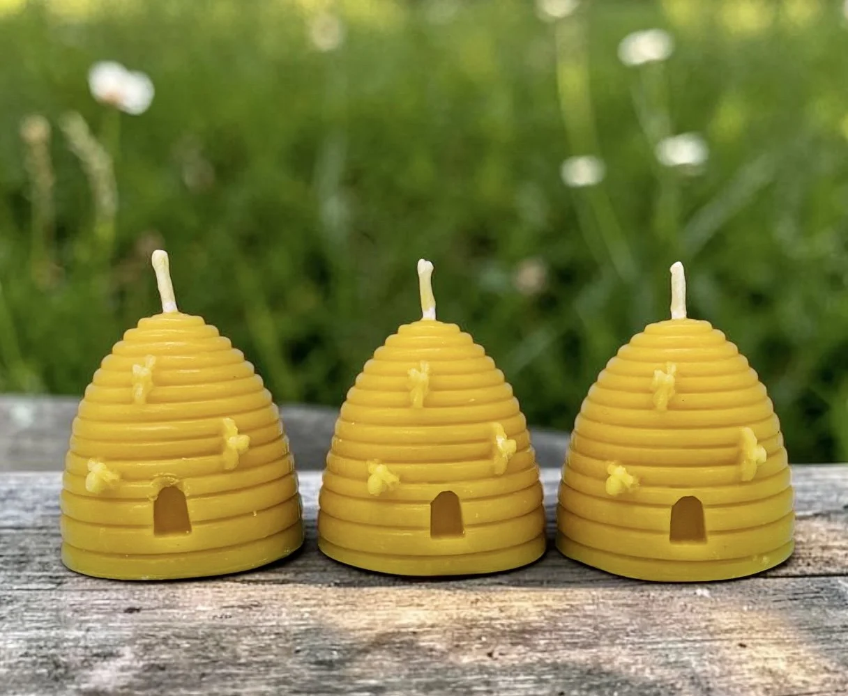 Three beeswax candles shaped like beehives with small door and honeycomb details, placed on a wooden surface outdoors.