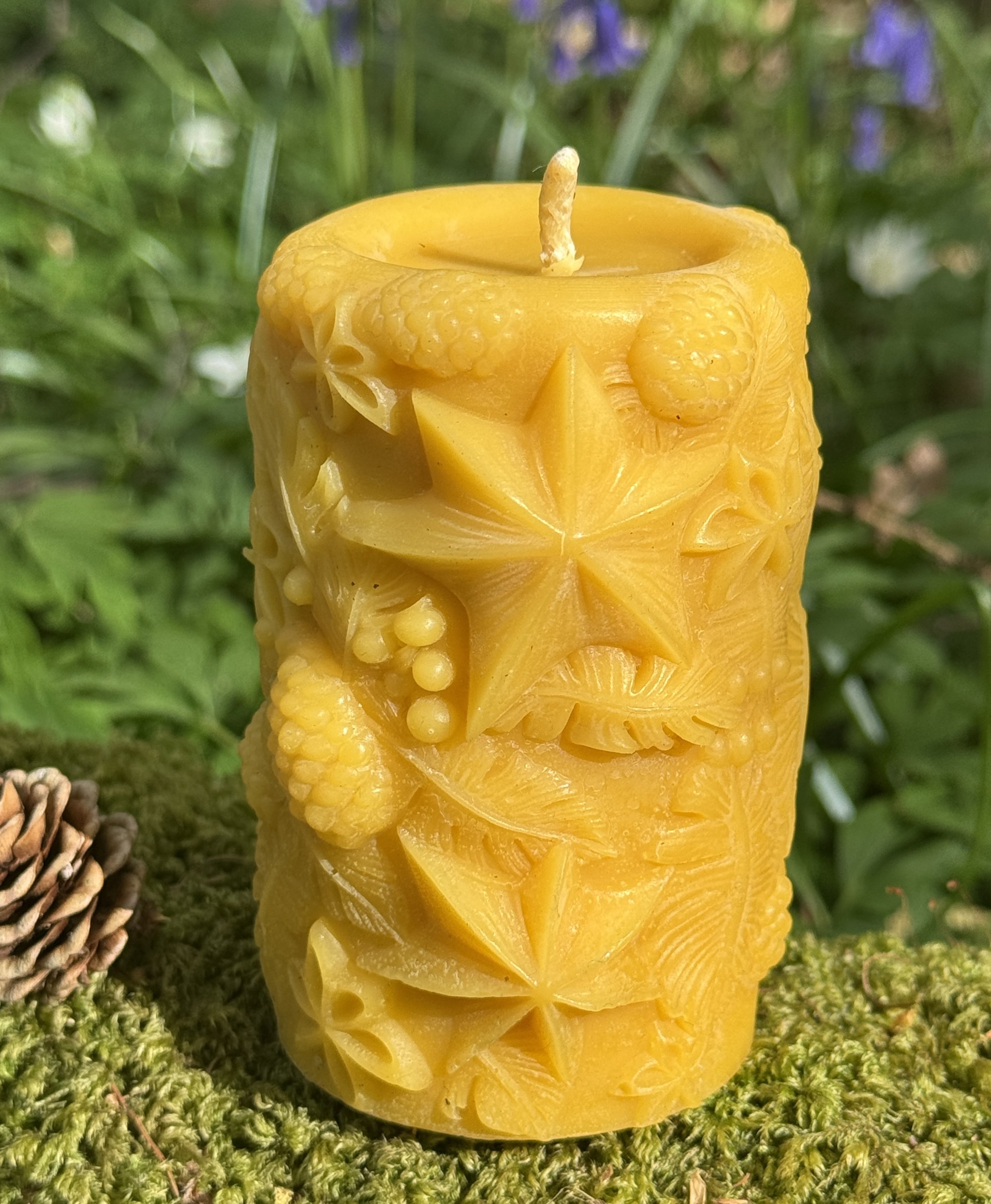 A carved yellow beeswax candle with intricate patterns and floral motifs, placed on green moss with a pinecone nearby, outdoors.