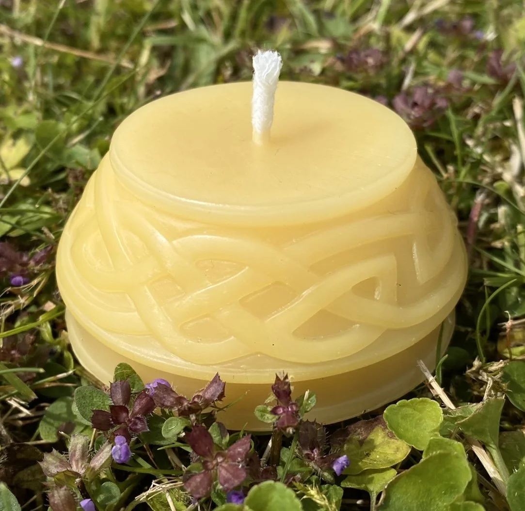 A pure beeswax Celtic Knot candle sitting on grass lawn with small flowers.