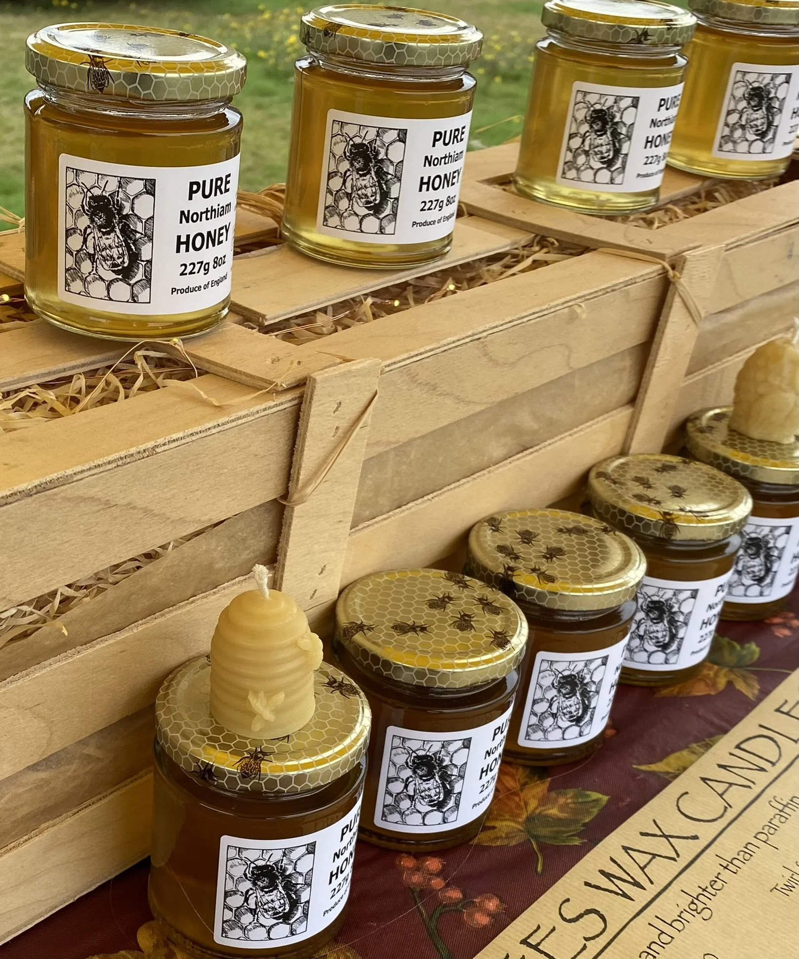 Close-up of jars of honey on a wooden display shelf at an outdoor market, with a background of grass and flowers.