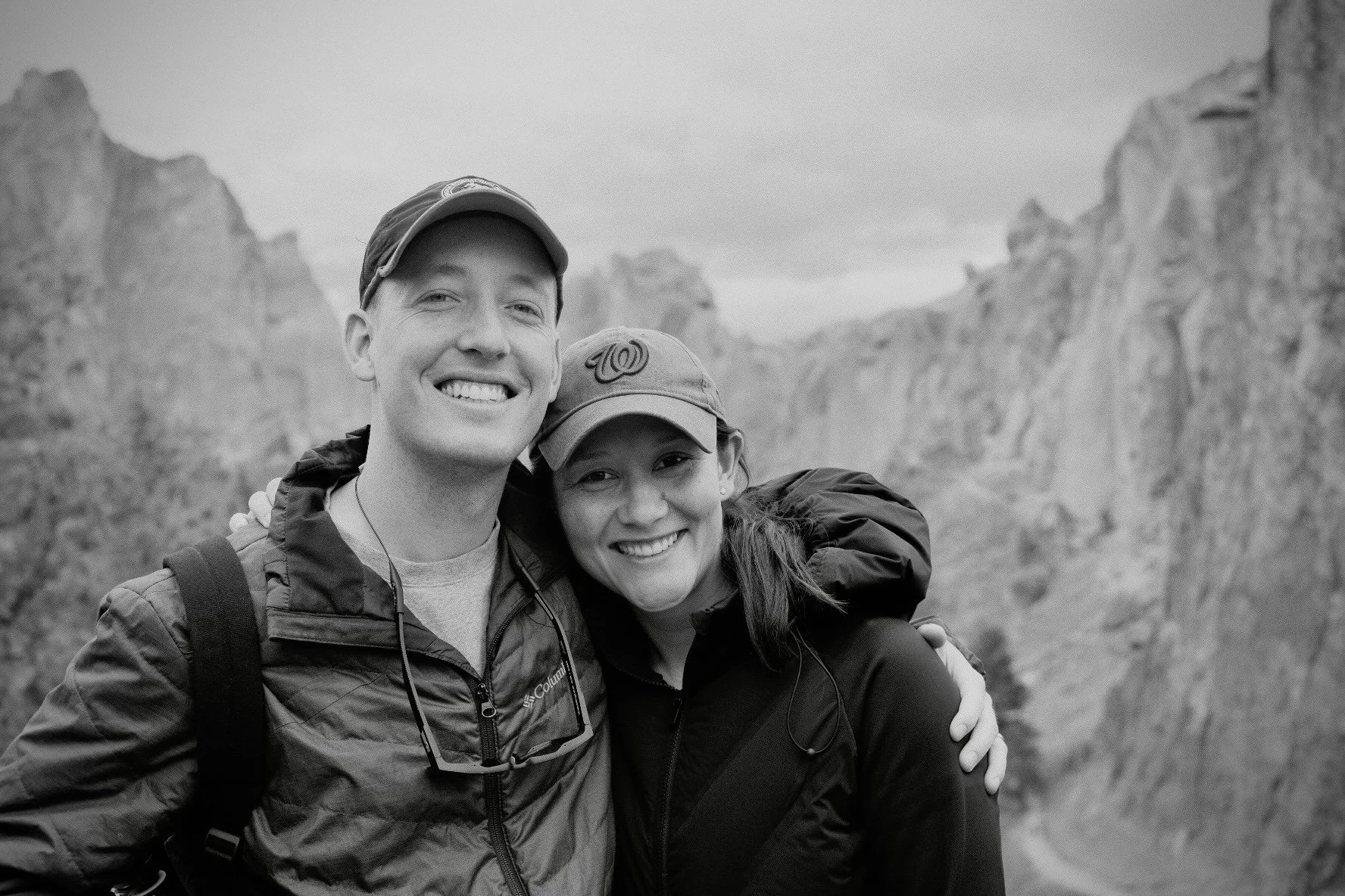 A smiling man and woman with their arms around each other, taking a selfie outdoors with mountains in the background.