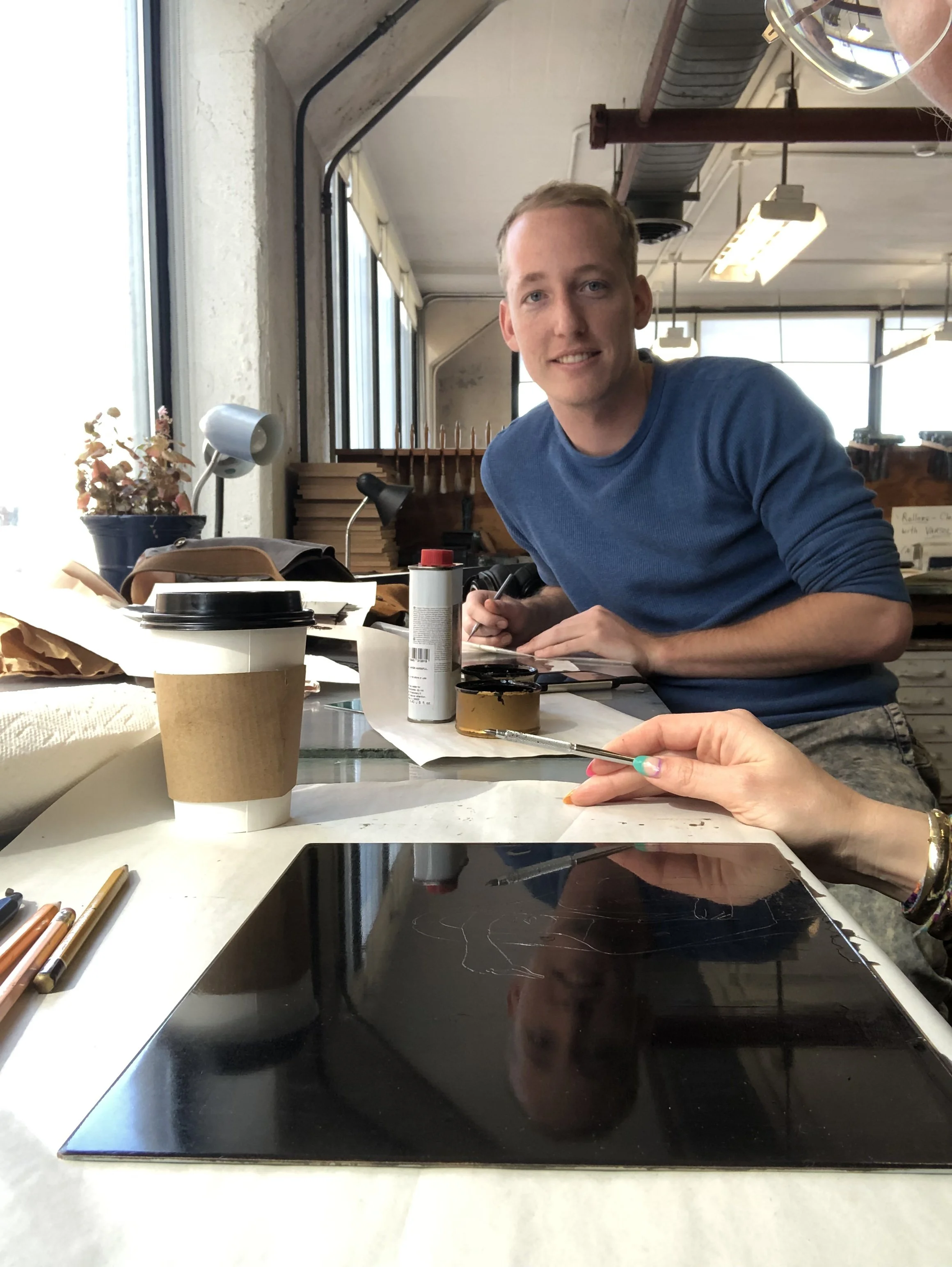 A picture of the artist Will, sitting at a cluttered table in a bright, industrial-style room with large windows, writing with a pen. The table has various items including coffee cups, a black tray, and colored pencils.
