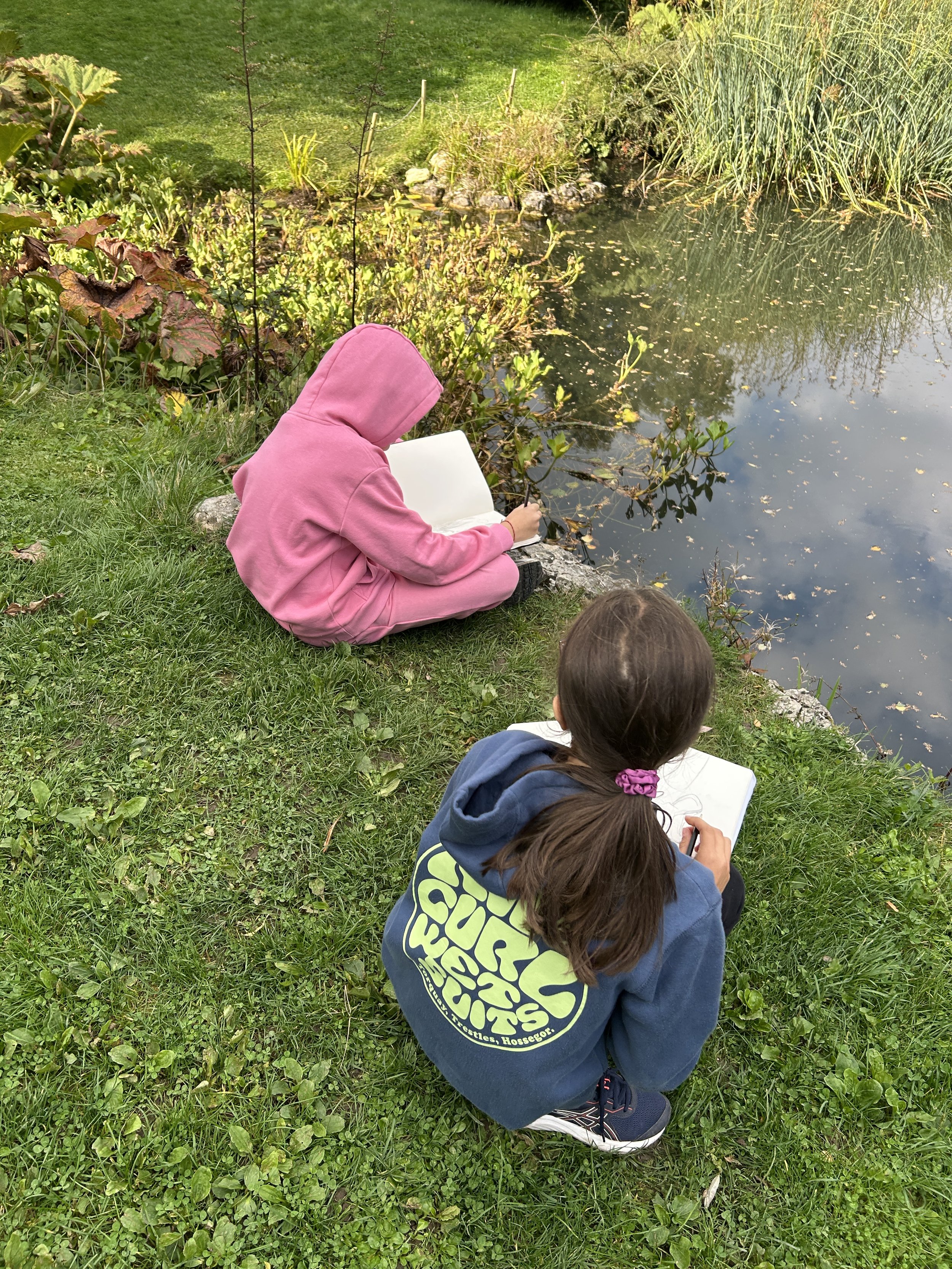 Two children sitting on grass near a pond, drawing in sketchbooks. One child wears a pink hoodie, the other a blue hoodie with yellow lettering on the back.