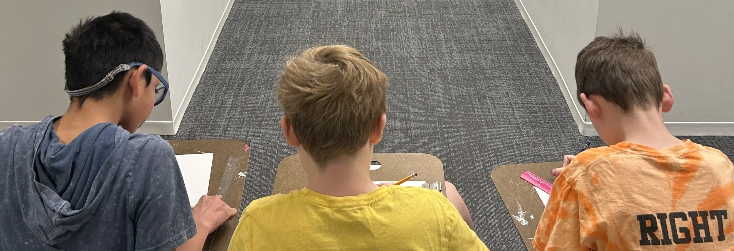 Three boys sitting at desks, viewed from behind, working on assignments in a classroom.