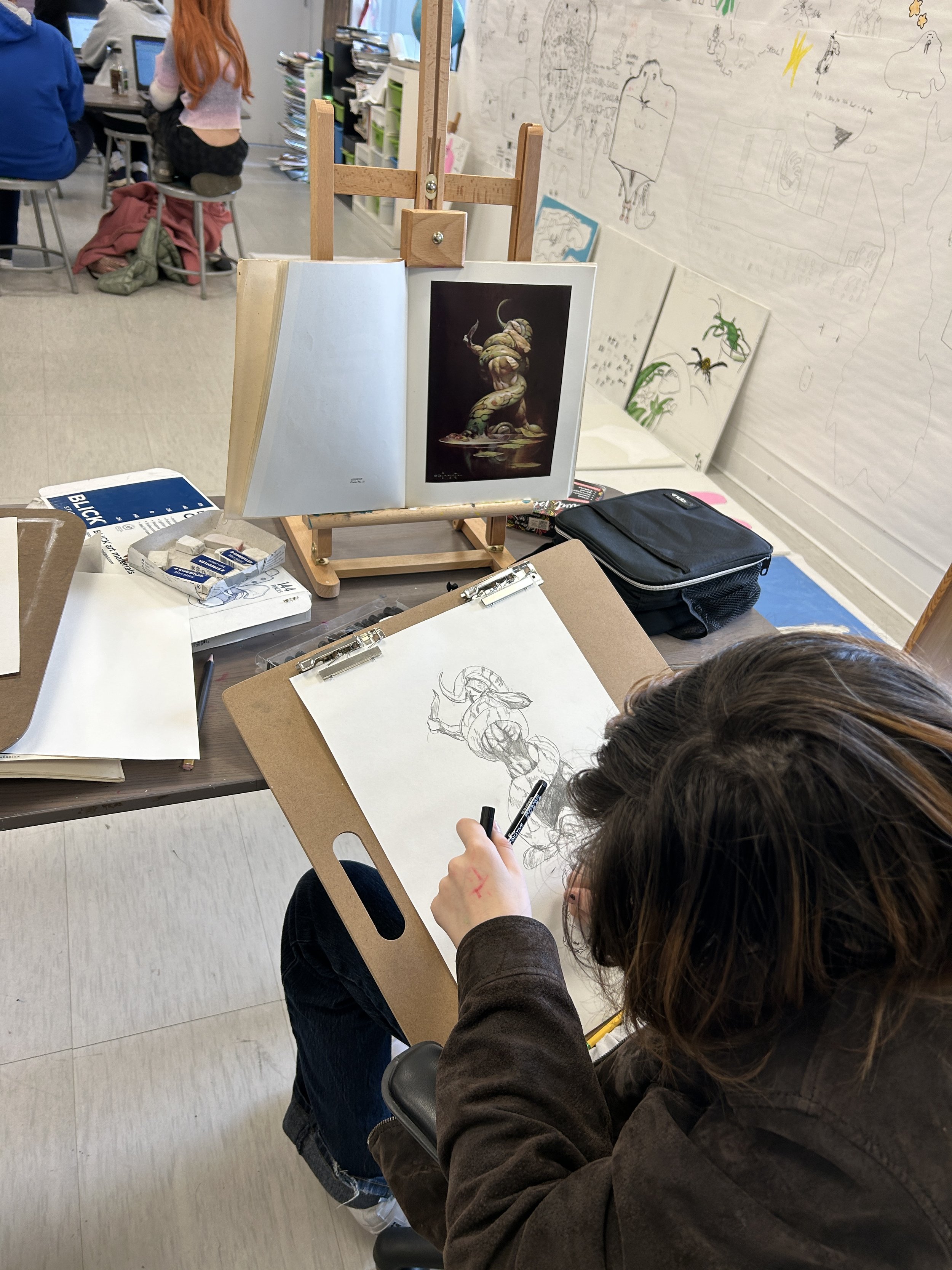 A person drawing a fantasy creature on a sketchpad, with an open art book displaying a 3D creature sculpture propped up on a wooden easel in front of the artist. There are other artworks on the wall behind the artist, and art supplies on the desk.