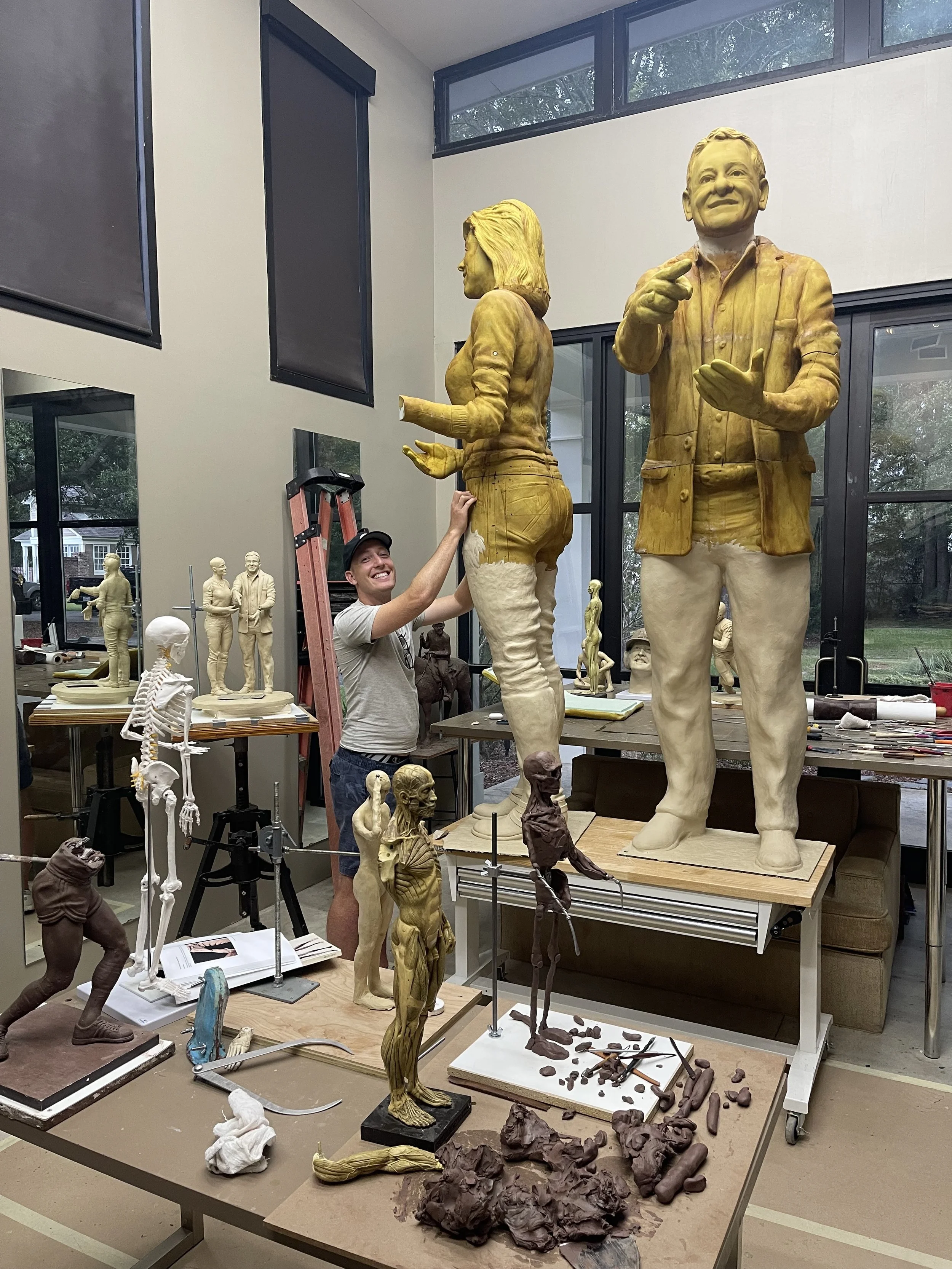 A man smiling and wearing a hat, standing in a sculpture workshop surrounded by unfinished sculptures and art supplies. He is touching one of the large wooden statues of a man and woman.