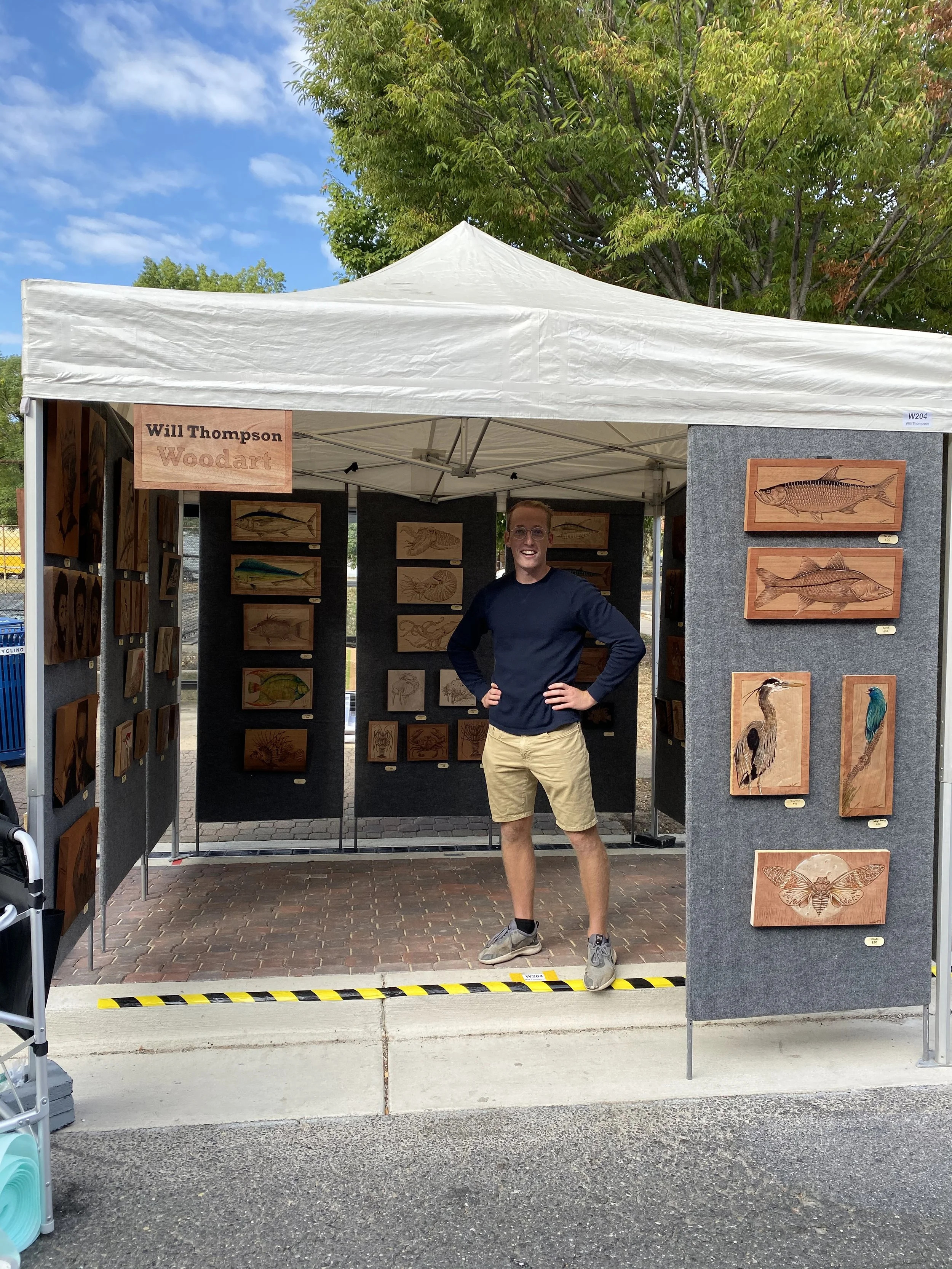 A man standing inside an art display booth under a white canopy, with art of fish, insects, and other animals mounted on foam boards displayed on the walls, and a sign with the name Will Thompson Woodart.