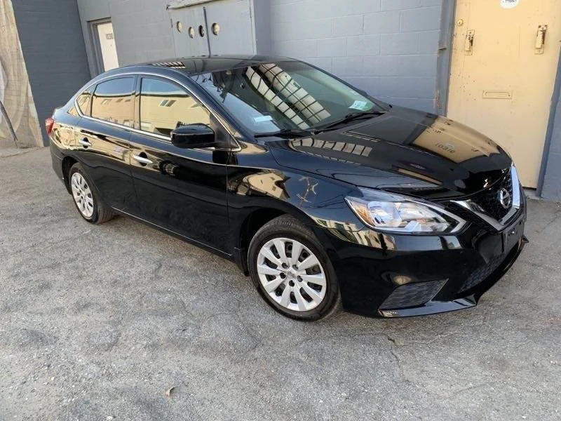 A shiny black sedan parked on a gravel ground near a building with a gray wall and yellow doors.