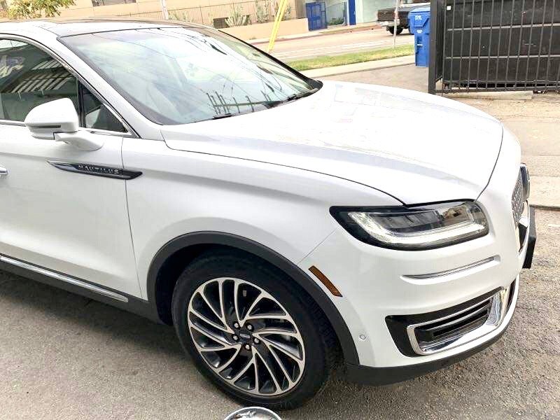 Front and side view of a white Audi vehicle parked on the street with a black fence and blue recycling bins in the background.