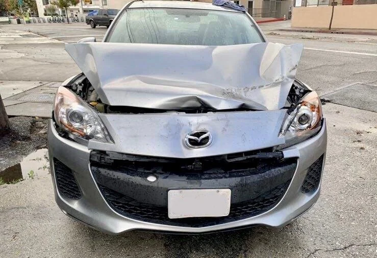 Silver Mazda car with front-end damage, crumpled hood, and broken bumper parked on city street