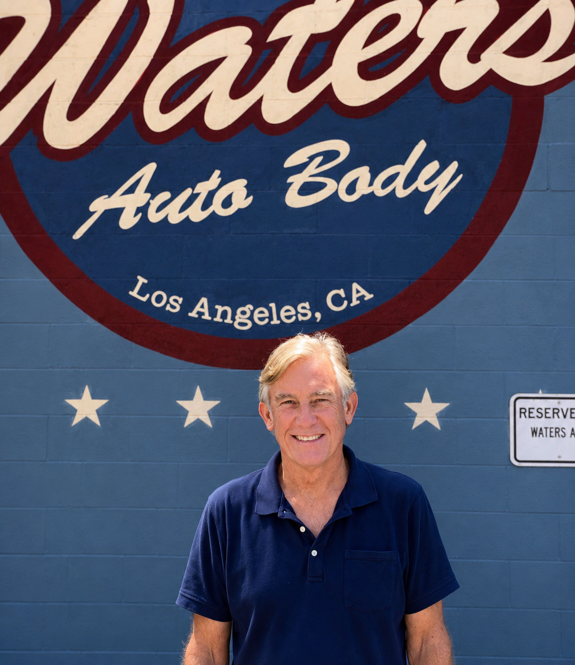 A smiling man with gray hair wearing a navy polo shirt standing in front of a large sign that says 'Waters Auto Body Los Angeles, CA' on a blue brick wall.