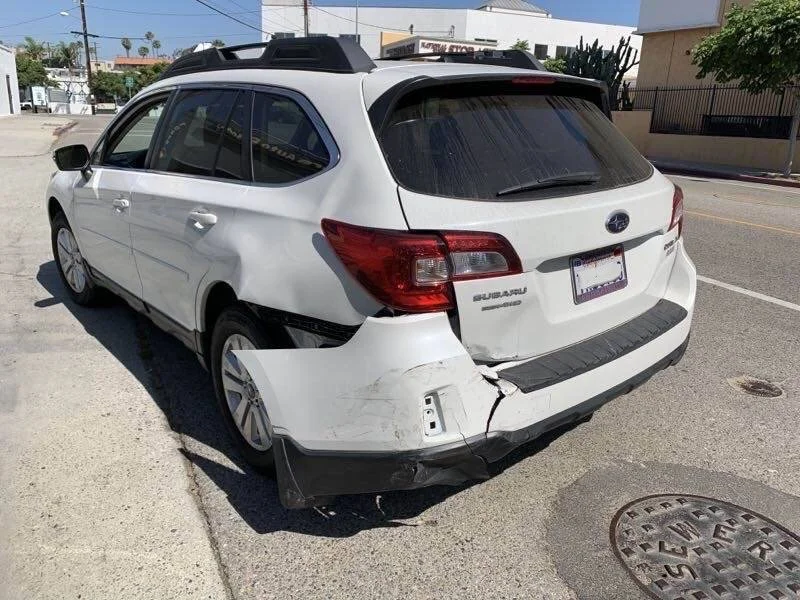 White Subaru Outback with rear damage and dents on the left side, parked on the street.