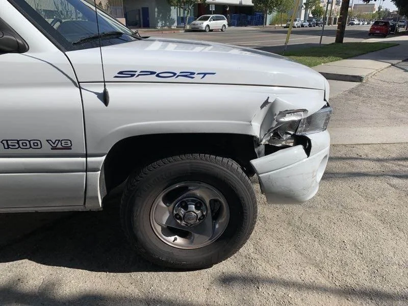 Silver pickup truck with a V8 engine badge and sports decal has front-end damage, including a crumpled bumper and damaged headlight, parked on a city street.
