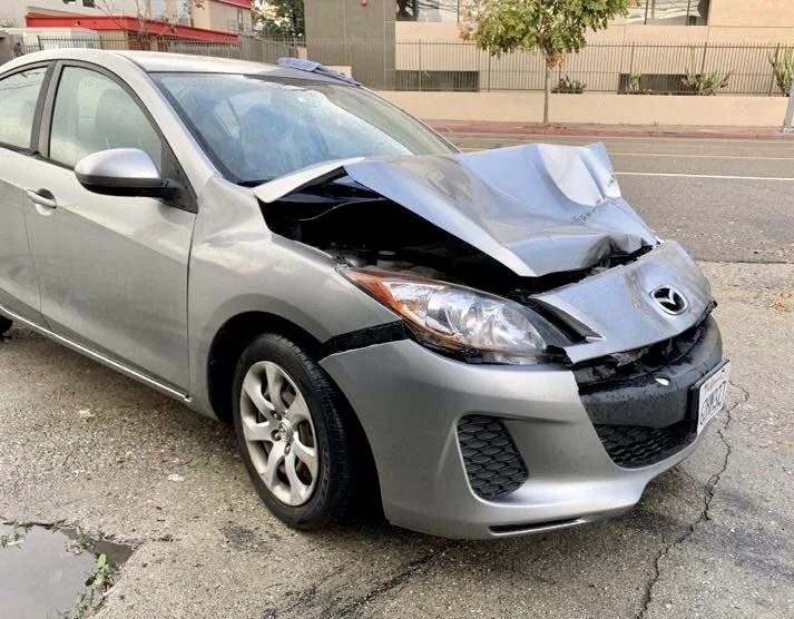 Gray sedan with front-end damage and a crumpled hood, parked on the side of a street.