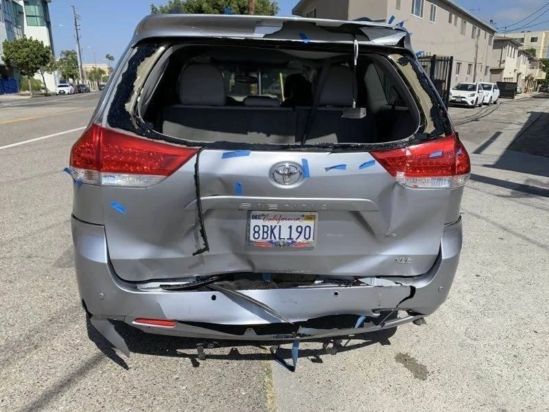 Damaged silver Toyota SUV with a shattered rear window, dented back, and a missing rear bumper, parked on a street near residential buildings.