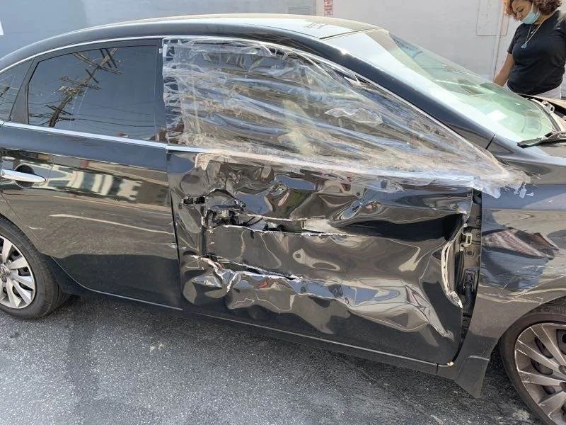 Damaged black car with significant side collision impact on the driver's side, missing front door, and shattered window, with a person wearing a face mask standing nearby.